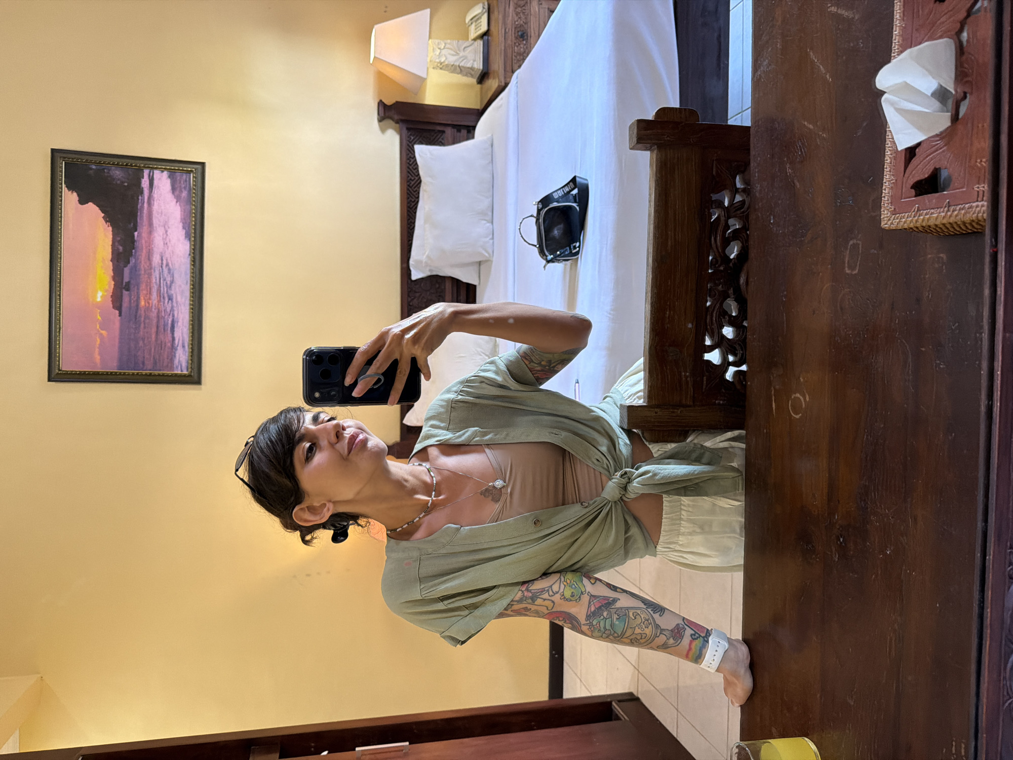 A woman is taking a selfie in a bedroom. She is wearing a light green button-down shirt tied at the waist over a beige tank top and off-white shorts. She has tattoos on her left arm and a necklace with a pendant. The bedroom has yellow walls, a bed with white linens, a wooden headboard, and a nightstand with a lamp and a phone. A black handbag is on the bed. A painting of a sunset over the ocean hangs on the wall. A wooden desk is in the foreground with a glass of orange juice and a tissue box. The scene is likely a hotel room in Denpasar, Indonesia.