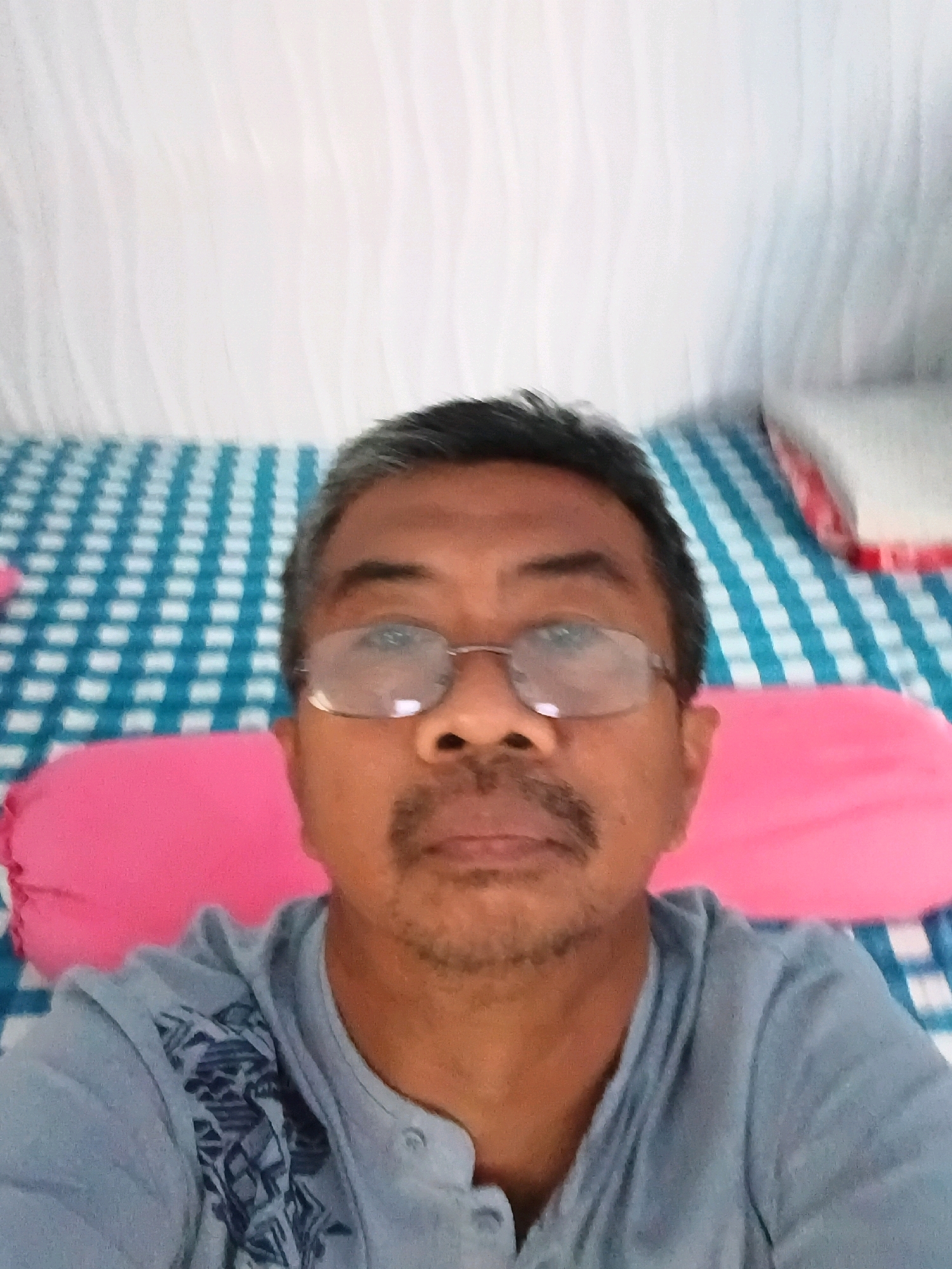 A selfie of a middle-aged man wearing glasses and a light blue shirt is captured against a blurred background of white and blue checkered fabric and pink pillows. The man has short, dark hair, a mustache, and stubble. He is looking directly at the camera. The scene appears to be indoors, likely a bedroom, based on the bedding and pillows. The image lacks any identifiable landmarks or context that would suggest a specific location within Tanggul Wetan, Indonesia.