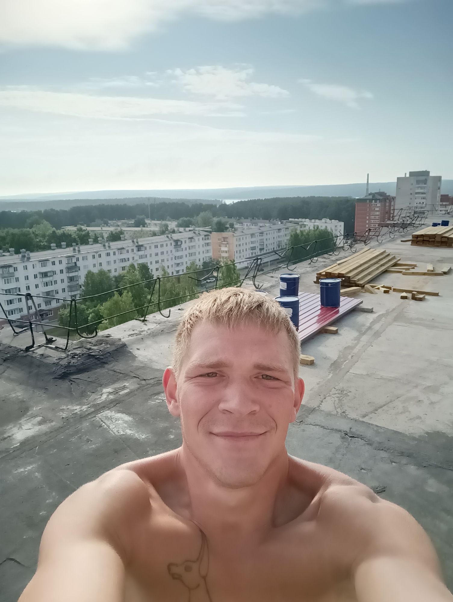 A shirtless man with blond hair and a tattoo on his chest smiles at the camera, taking a selfie from what appears to be a rooftop construction site. In the background, a city skyline is visible with apartment buildings, trees, and a hazy sky. Construction materials, including stacks of lumber and blue barrels, are present on the rooftop.