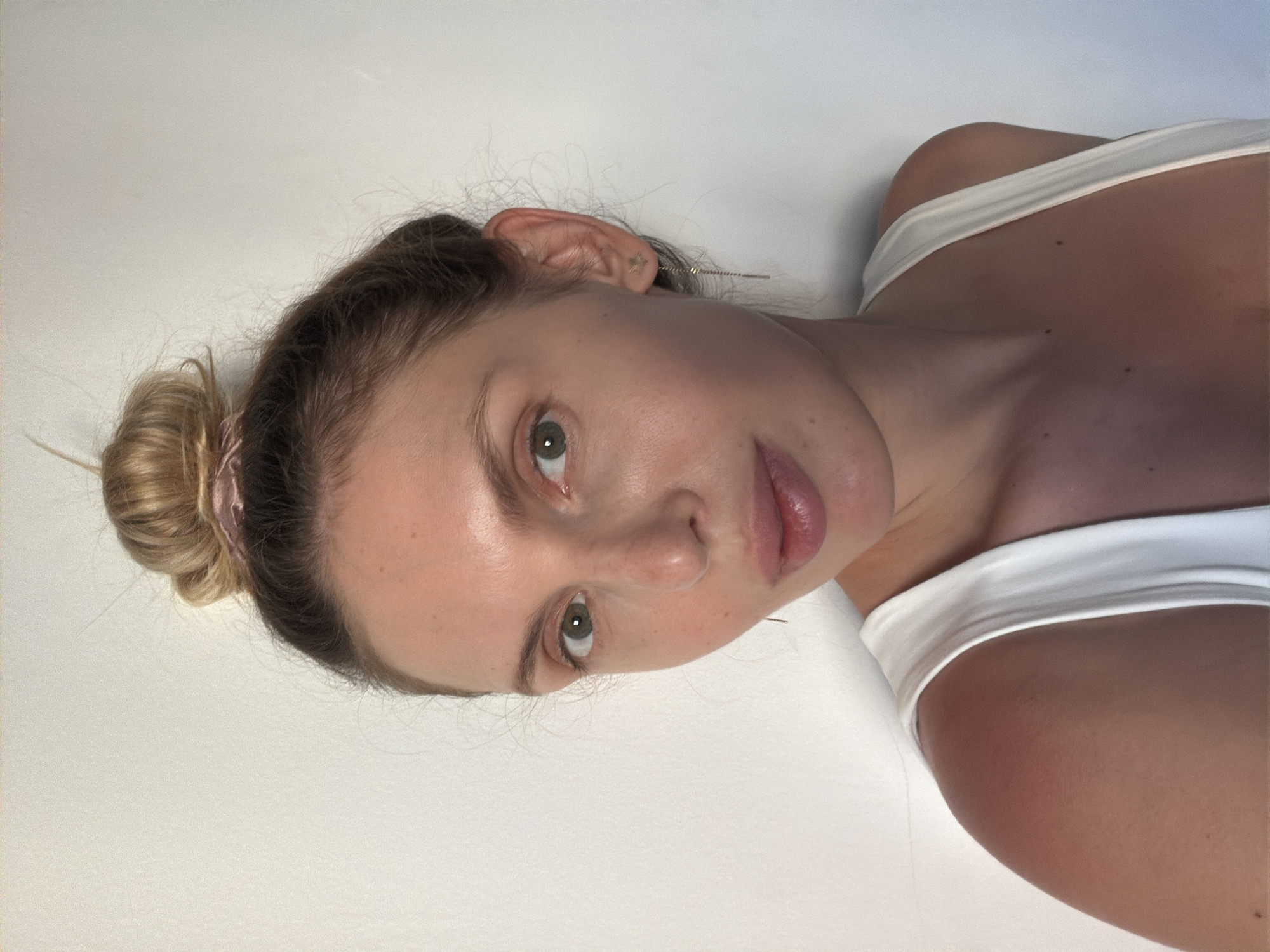 A young woman is photographed indoors against a white background. She has light skin, green eyes, and her hair is styled in a bun with a pink hair tie. She is wearing a white tank top. The location of Budva, Montenegro is not discernible from the image.