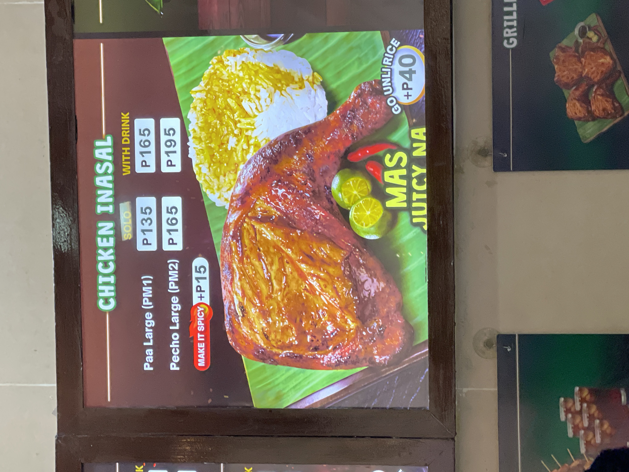 The image displays a digital menu board for "CHICKEN INASAL" in a casual restaurant setting, located in Davao City, Philippines. The menu lists two main items: "Paa Large (PM1)" (chicken leg/thigh quarter) available for P135 (Solo) or P165 (With Drink), and "Pecho Large (PM2)" (chicken breast) priced at P165 (Solo) or P195 (With Drink). An option to "MAKE IT SPICY" is available for an additional P15, indicated by a red button with a flame icon.
The central visual features a large, glistening grilled chicken leg quarter presented on a vibrant green banana leaf. Alongside the chicken is a mound of yellow-tinted rice, two halved calamansi limes, two small red chili peppers, and a small, dark dipping sauce container partially visible on the right. Text overlaid on the image reads "MAS JUTCY NA" in yellow, translating to "More Juicy Now." Another option, "GO UNLI RICE +P40," is displayed on a curved banner, offering unlimited rice for an additional 40 Philippine Pesos.
The menu board is enclosed within a dark brown wooden frame and is mounted against a light beige or off-white wall. Below the main menu, a partial section of another menu board is visible, featuring a dark green background with "- GRILLE" as a partial title, and an image of several pieces of grilled meat (likely pork) on a banana leaf with a small dipping bowl. Small, circular transparent suction cups are present on the wall, suggesting the attachment mechanism for the lower menu or other posters. No people are visible in the scene.