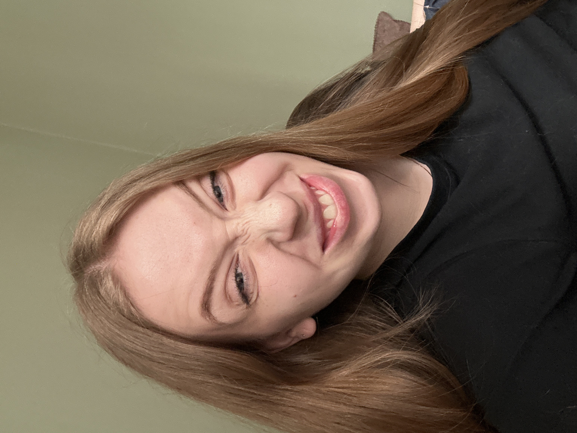 A young woman with long, light brown hair is shown in a close-up, tilted shot. She is wearing a black shirt. Her face is scrunched up in a grimace, with her eyes squinted and mouth showing her teeth. The background is a plain green wall. There is nothing indicating the location is in the city of Chaikovsky, Russia.