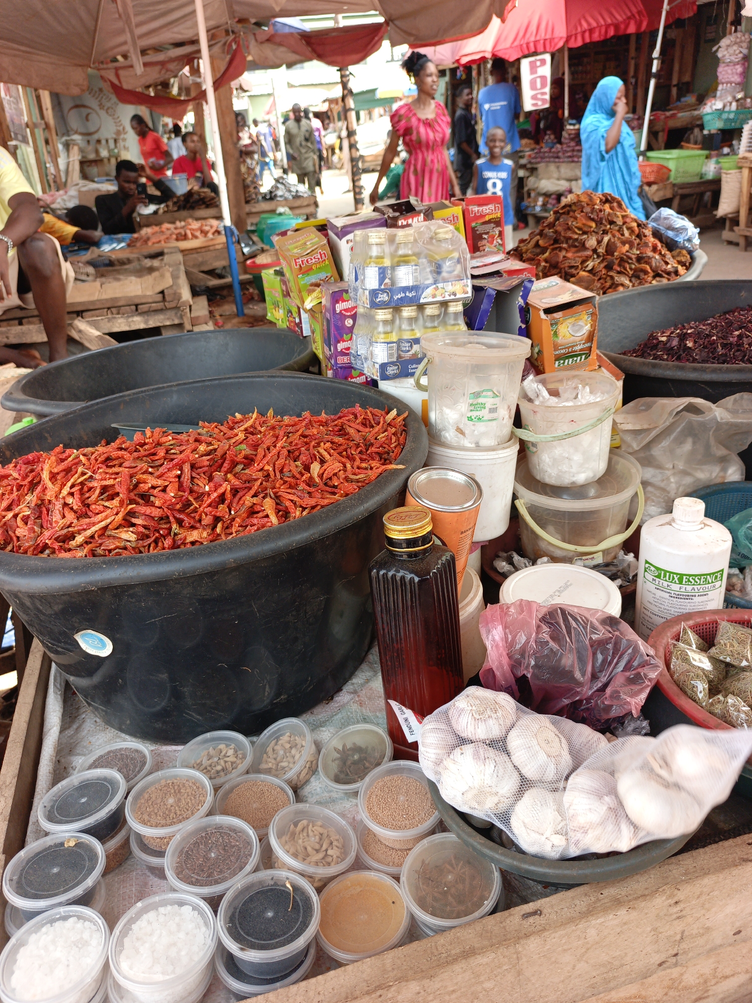 This is a bustling outdoor market scene likely located in Akwanga, Nigeria. The foreground is dominated by a vendor's stall filled with various food items, spices, and condiments. A large black tub overflows with dried red chili peppers. Nearby are containers of various spices, including salt, black peppercorns, mustard seeds, and others. There are also containers and bottles of oil, flavorings, and seasonings. A bag of garlic is prominently displayed.
In the background, people are walking around, some wearing brightly colored clothing. There are other market stalls visible under red and white canopies.