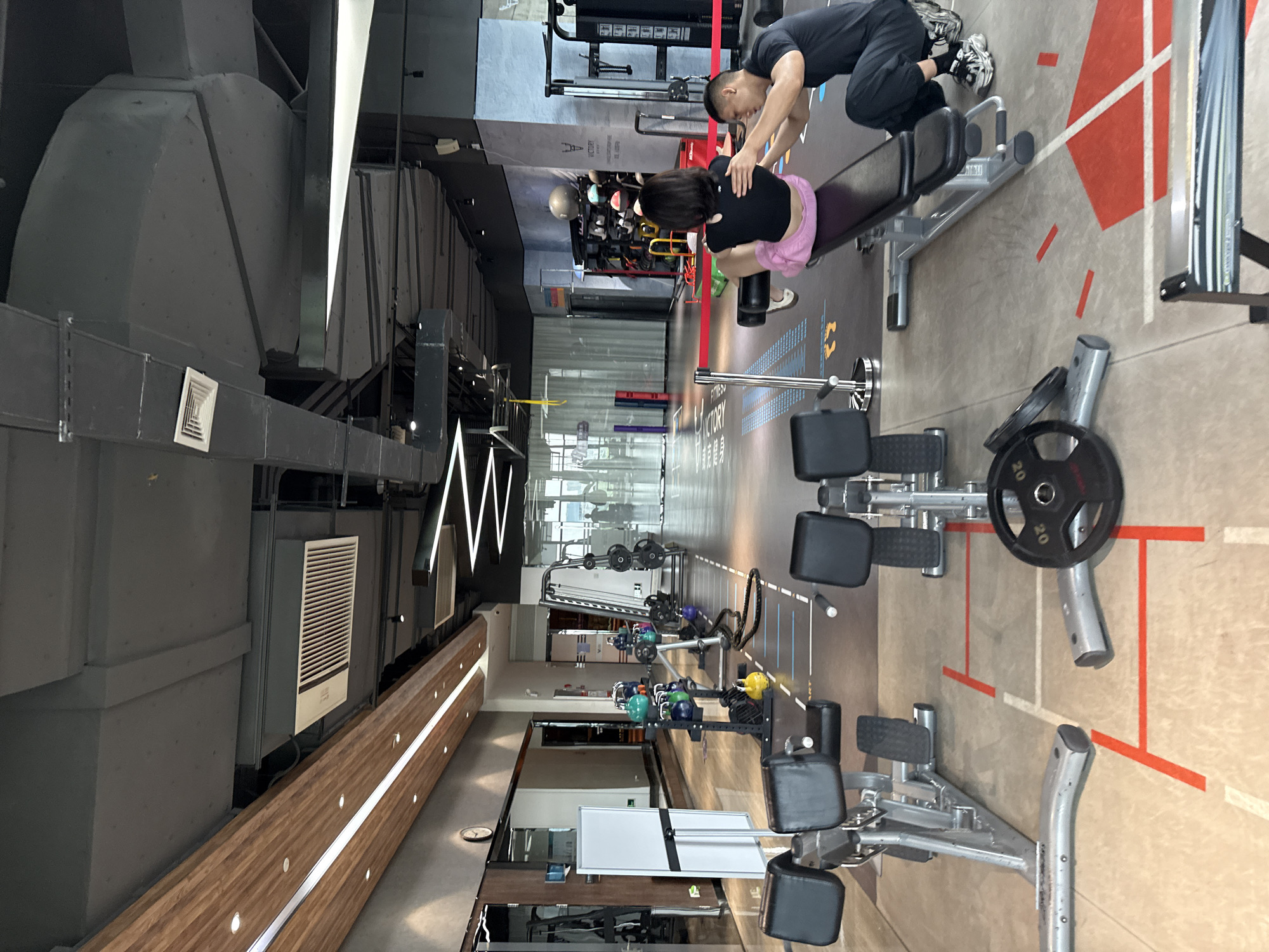 The image shows the interior of a modern gym located in Haizhu District, China. The gym features high ceilings with exposed ductwork and lighting fixtures. The floor is a light-colored concrete with various training markings, including red lines and white squares.
In the foreground, there are several pieces of weight training equipment. On the left, a silver and black weight bench with adjustable backrests is visible. To the right of this bench, a single 20-kilogram weight plate rests on the floor. Further back and to the right, another weight bench, also silver and black, is occupied by two individuals. A woman, wearing a black sports top and pink shorts, is seated on the bench, facing away from the camera, with her legs extended. A man, dressed in a black t-shirt and black pants, is standing behind her, with his hands on her lower back, appearing to assist her with an exercise.
In the middle ground of the gym, various fitness equipment is arranged. There is a rack of colorful kettlebells, a barbell with weights, and a rope. A cable crossover machine is visible further in the background. The gym also has mirrored walls on the left, reflecting some of the equipment.
In the background, through a large glass partition, another section of the gym is visible, with more machines and a few other individuals training. The overall environment suggests a well-equipped and organized fitness facility.