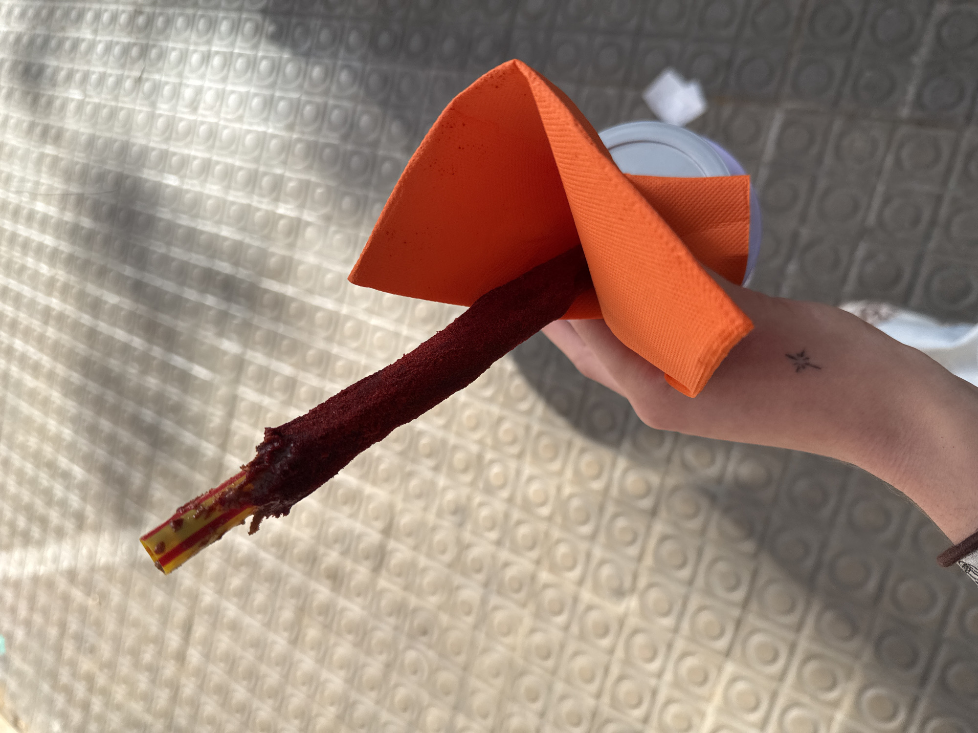 A person holds a tamarind candy stick, likely in Barcelona, Spain, due to the style of the street pavers in the background. The candy stick is red and coated in a granular substance, held in an orange napkin. It appears to be placed in a cup. The person's fair-skinned arm has a small tattoo on the wrist. The background shows an outdoor setting with sunlit pavers, casting shadows.
