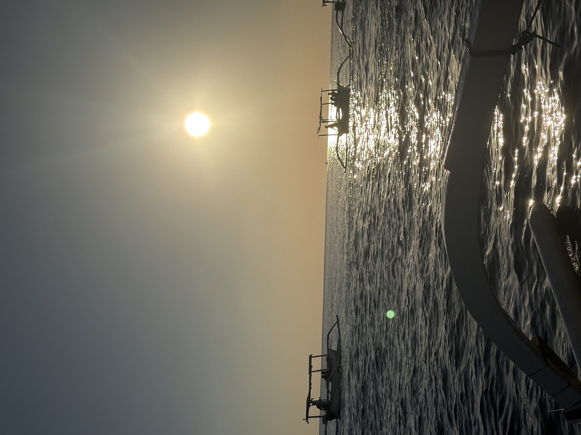 The image depicts a serene seascape at sunset, likely in or near Seririt, Indonesia. The sun, a bright orb, is positioned in the upper right quadrant of the sky, casting a warm, golden glow that reflects brilliantly on the rippling water. Several traditional Indonesian outrigger boats, known as "jukung," are visible. The closest boat in the foreground is partially captured, with its white outriggers and hull visible. Further in the distance, two other jukung boats are sailing. One boat on the left has silhouetted figures on board, and another boat to the right also appears to carry passengers. The ocean is a deep, calm blue, with gentle waves. The sky is a gradient of pale blue at the top, transitioning to a soft yellow and orange near the horizon, indicating the setting sun. A faint green circular light artifact is visible in the water in the middle left of the frame. The overall atmosphere is tranquil and picturesque, capturing a moment of calm on the sea.
