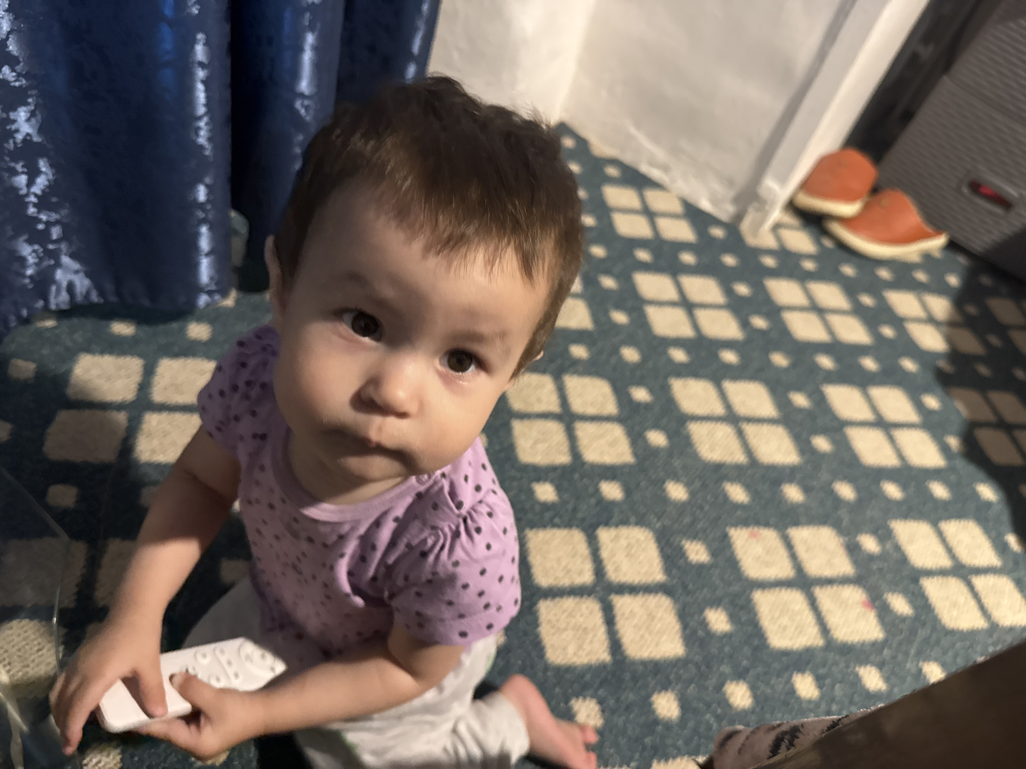 A close-up, high-angle shot captures a baby seated on a patterned blue and cream rug. The baby is wearing a purple t-shirt with small black polka dots and gray pants. The baby is holding a white object, possibly a remote control, in its hands. In the background, dark blue curtains are visible on the left, and a white object and a pair of orange slippers are on the right. The scene is indoors. The location of Chardara, Uzbekistan, is not evident from the image.