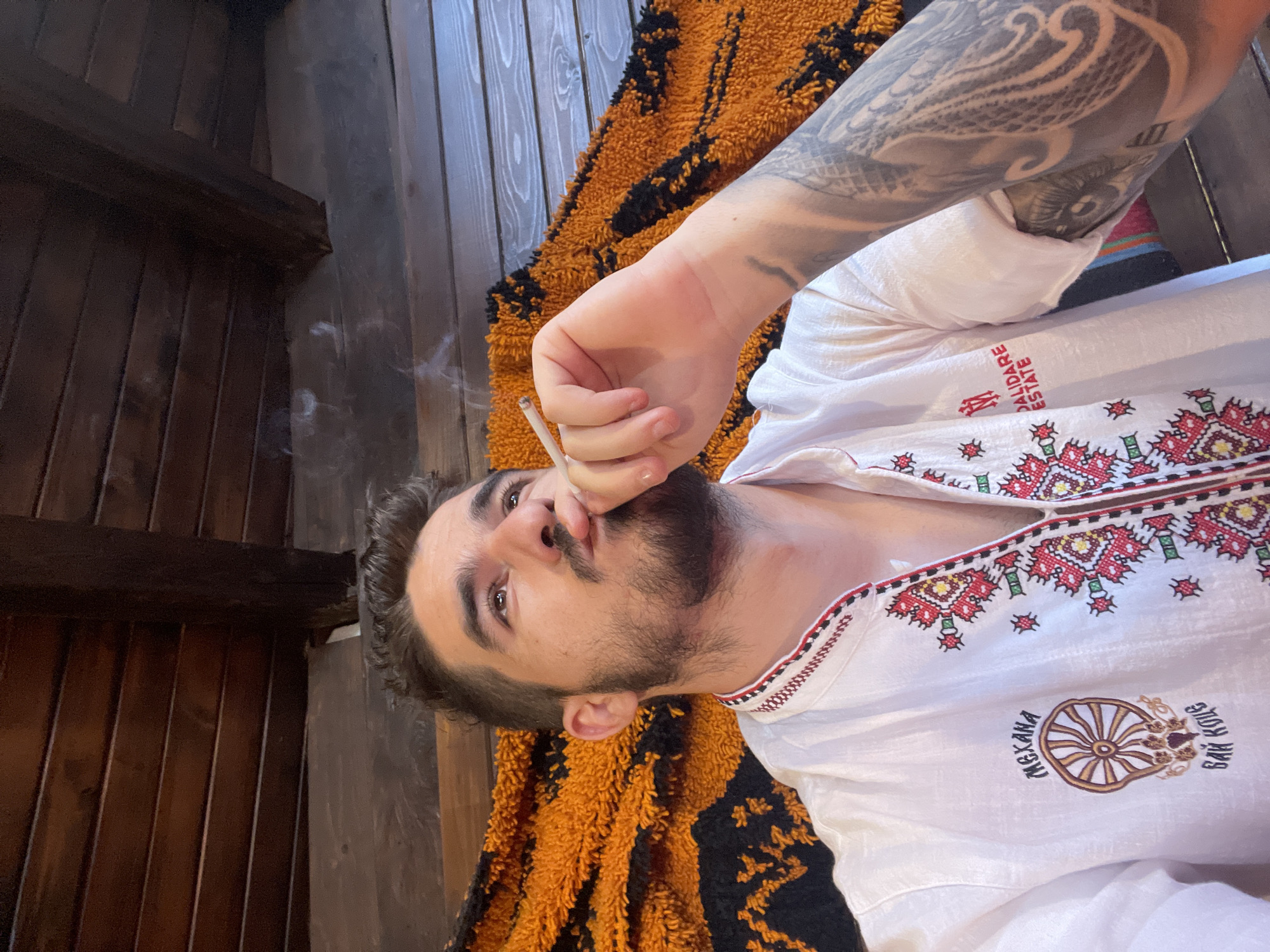 A man is shown from the chest up, holding a cigarette to his lips and exhaling smoke. He is looking up and to his left. He has dark hair, a beard, and is wearing a white shirt with red and black embroidery. Behind him is an orange and black textured fabric, and wooden planks form the ceiling. A tattoo is visible on his left arm. The scene is indoors, under a wooden ceiling.