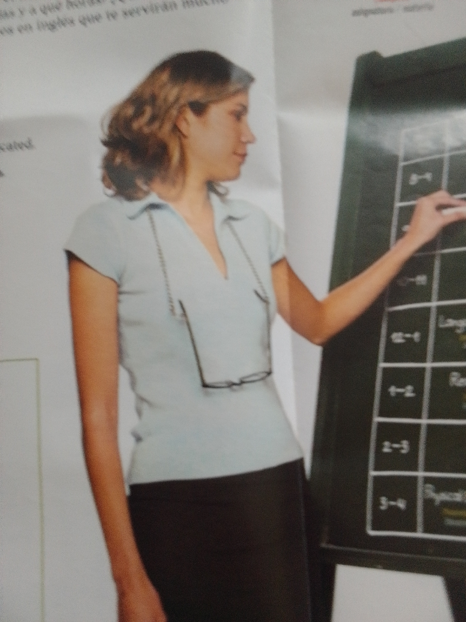 The image shows a woman standing in front of a chalkboard or a similar teaching board. She is wearing a light blue polo shirt and a dark skirt. Glasses are hanging around her neck. The chalkboard has a grid with some writing and numbers, suggesting it's used for teaching or organization. The woman's right hand is near the board, and she appears to be pointing or interacting with it. The overall scene suggests a classroom or a learning environment. There are some faint words visible on the left side of the image, and some text on the chalkboard. The quality of the image is somewhat blurry, making it difficult to read all the text clearly.