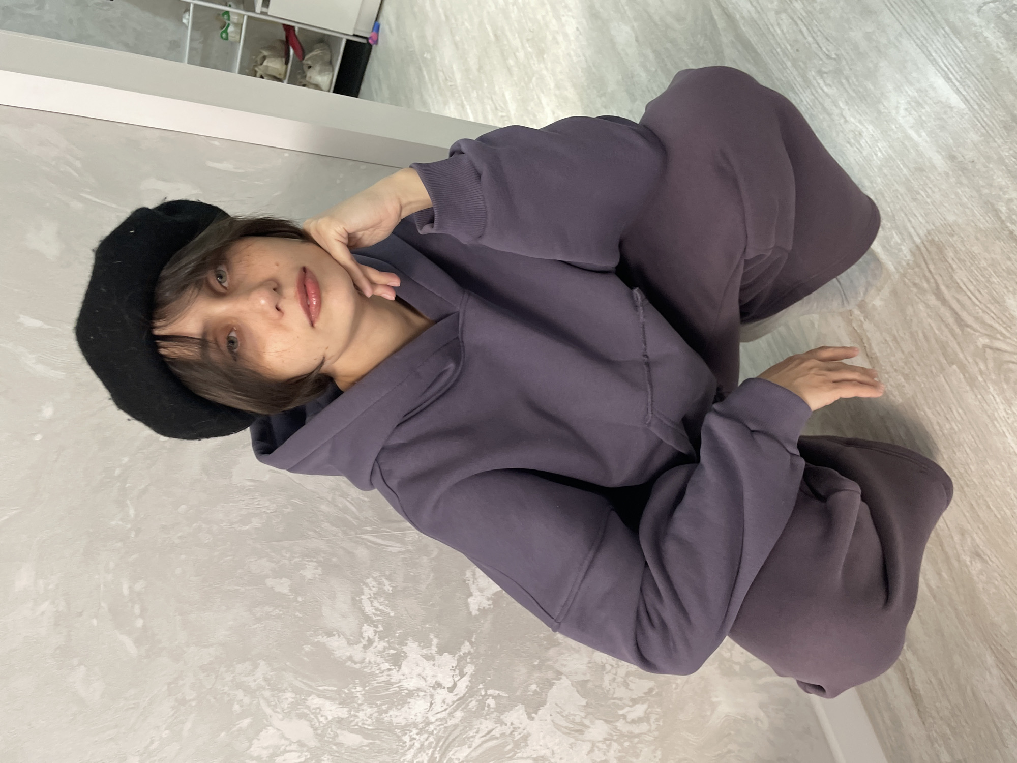 A young woman with fair skin, short brown hair, and light-colored eyes is sitting on a light-colored, patterned floor. She is wearing a black beret and a purple hoodie and sweatpants set. She is facing the camera with her head tilted slightly. Her left hand is touching her face. The background includes a white wall and a glimpse of shelving in the upper left corner. There is no indication of location in Izhevsk, Russia, or any other city.