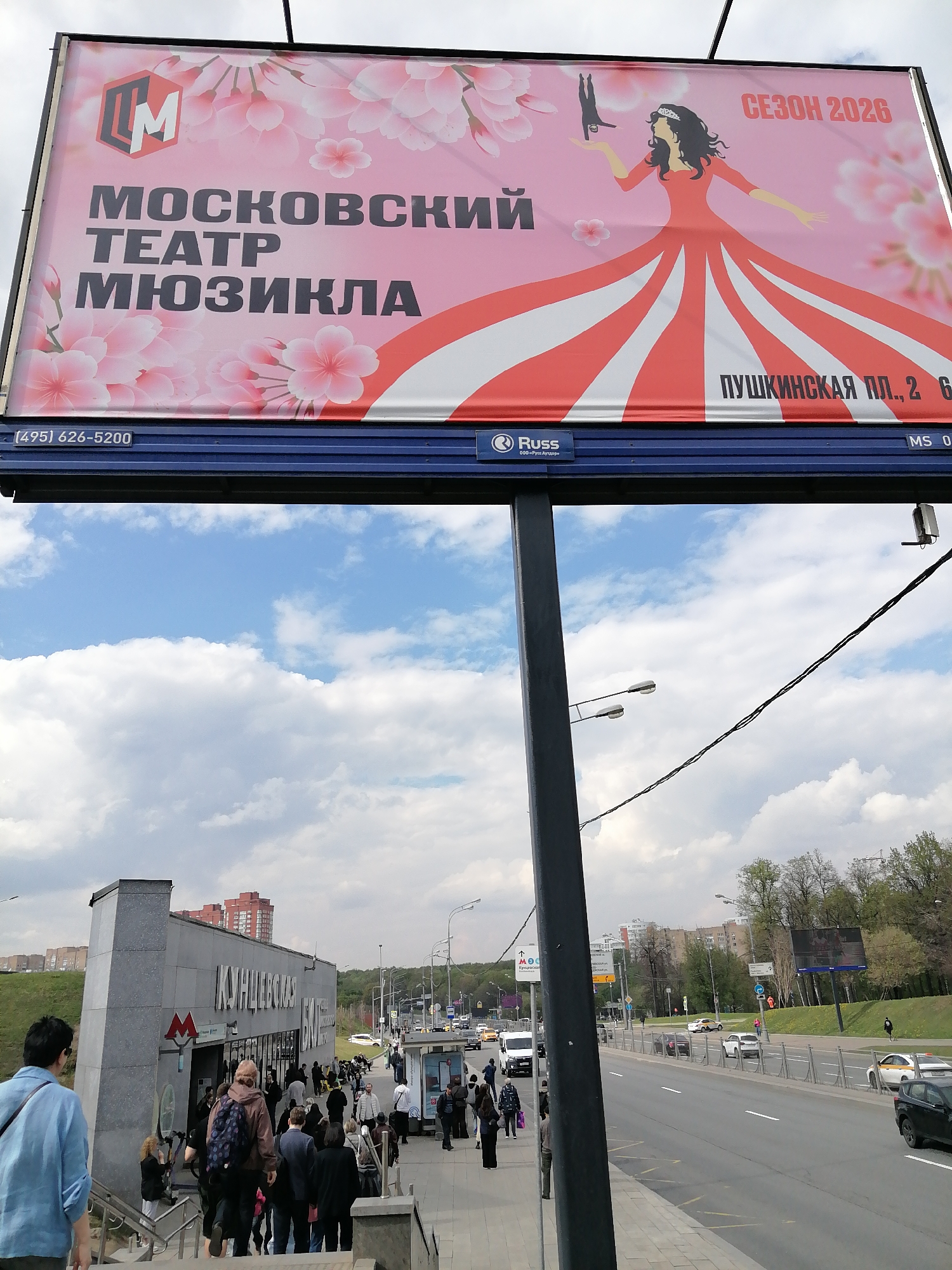 Here is a detailed description of the image:
**Overview**
The image depicts an outdoor scene, primarily focused on a large advertising billboard and a nearby subway entrance. The setting appears to be an urban area.
**Details**
* **Billboard:**
* Located at the top of the frame, advertising the "Moscow Musical Theater."
* Features the theater's logo, the name of the theater in Russian, a cartoon woman holding a puppet.
* Advertises the theater season as "2026"
* Includes a phone number.
* **Subway Entrance:**
* A modern-style subway entrance is visible on the left side of the image.
* The entrance has the word "Kuntsevskaya" written above in Russian.
* People are seen entering and exiting the subway.
* **People:**
* Numerous people are present, some walking towards the subway, others standing around.
* **Street:**
* A street runs along the right side of the image, with cars and other vehicles visible.
* The street has pedestrian barriers.
* **Environment:**
* The sky is partly cloudy.
* Buildings and greenery can be seen in the background.
* **Objects:**
* Light poles are present.
* Power lines run across the image.
* A trash bin is visible.
**Inferences**
* The image appears to be taken in Moscow, Russia. This is suggested by the text in Russian.
* The people in the scene are likely commuters and travelers.
* The presence of the billboard and subway station indicates a busy, urban environment.