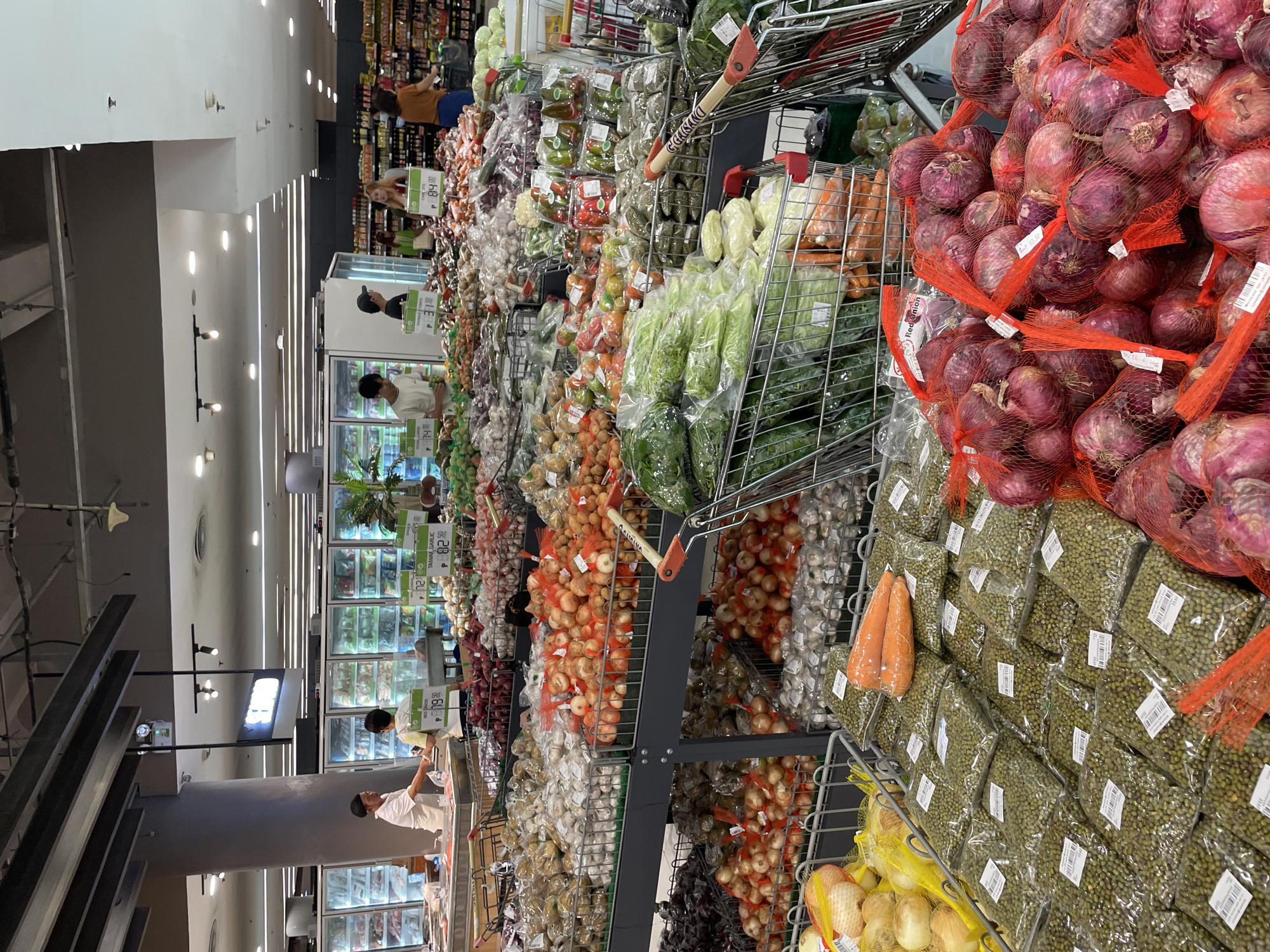 A wide shot captures a bustling produce section of a supermarket in Davao City, Philippines. Various fruits and vegetables are displayed in metal carts and on shelves. In the foreground, bags of mung beans and red onions are prominently featured, with two carrots placed on top of the mung bean bags. Behind these, an assortment of produce including leafy greens, tomatoes, bell peppers, and more onions are visible in shopping carts. In the midground, staff members are seen interacting with customers near refrigerated displays. The background shows aisles of food products and additional shoppers browsing the market. The lighting suggests an indoor, well-lit environment typical of a supermarket.