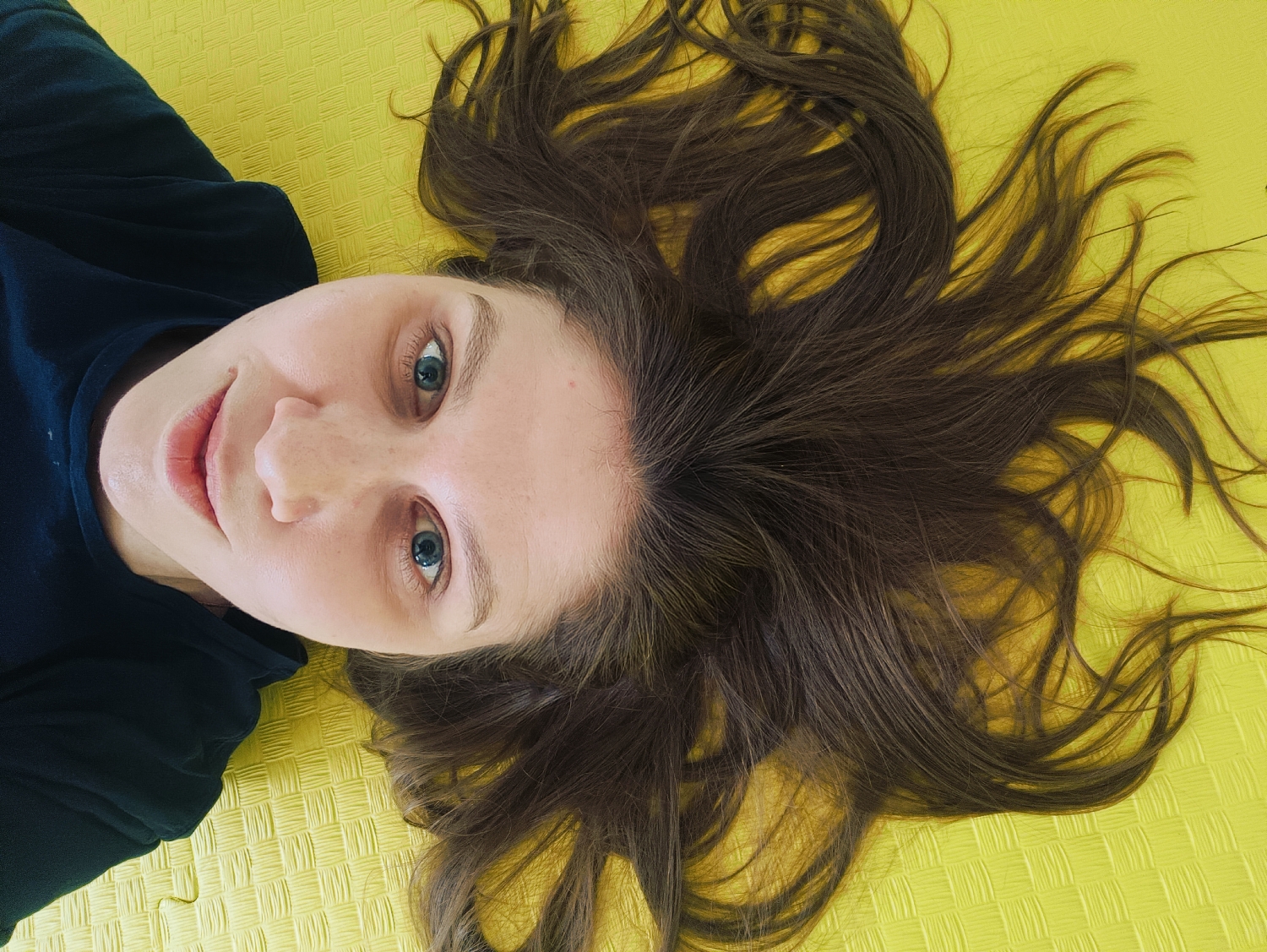 The image displays a person, likely a woman, lying on her back on a vibrant yellow, textured surface. The perspective is from directly above, capturing her head and upper torso. She has fair skin, blue eyes, and long, dark brown hair that is spread out in a wide fan around her head, covering a significant portion of the bright yellow background.
She is wearing a dark, possibly black, long-sleeved shirt. Her facial expression is relaxed and calm, with a slight, gentle smile on her lips, and her blue eyes are open and looking directly towards the camera. Her skin appears natural with some subtle freckles visible.
The yellow surface beneath her has a distinct, repeating woven or grid-like pattern, suggesting it could be a type of mat, such as a yoga mat or a soft play mat. The lighting in the image is bright and even, highlighting the details of her face and hair, as well as the texture of the background. There is no discernible text visible in the image. The overall scene conveys a sense of peacefulness and direct eye contact.