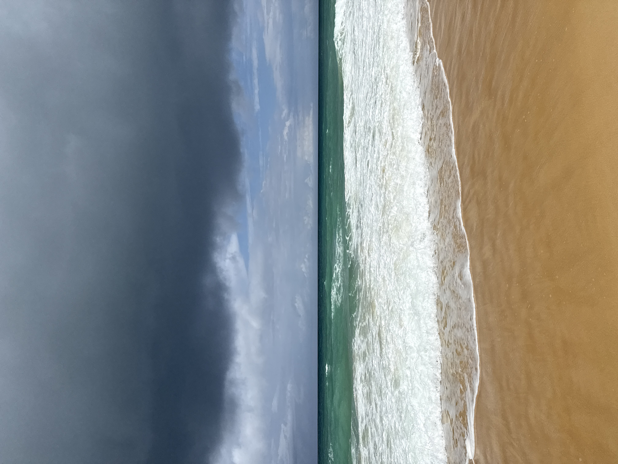 The image shows a beach scene in Karon, Thailand. The foreground is a sandy beach with a wave lapping at the shore. The ocean is a turquoise green with white foam from the waves. In the background, there are dark, heavy storm clouds gathering in the sky, with some lighter clouds and a sliver of blue sky visible. There are no people or man-made objects visible in the image.