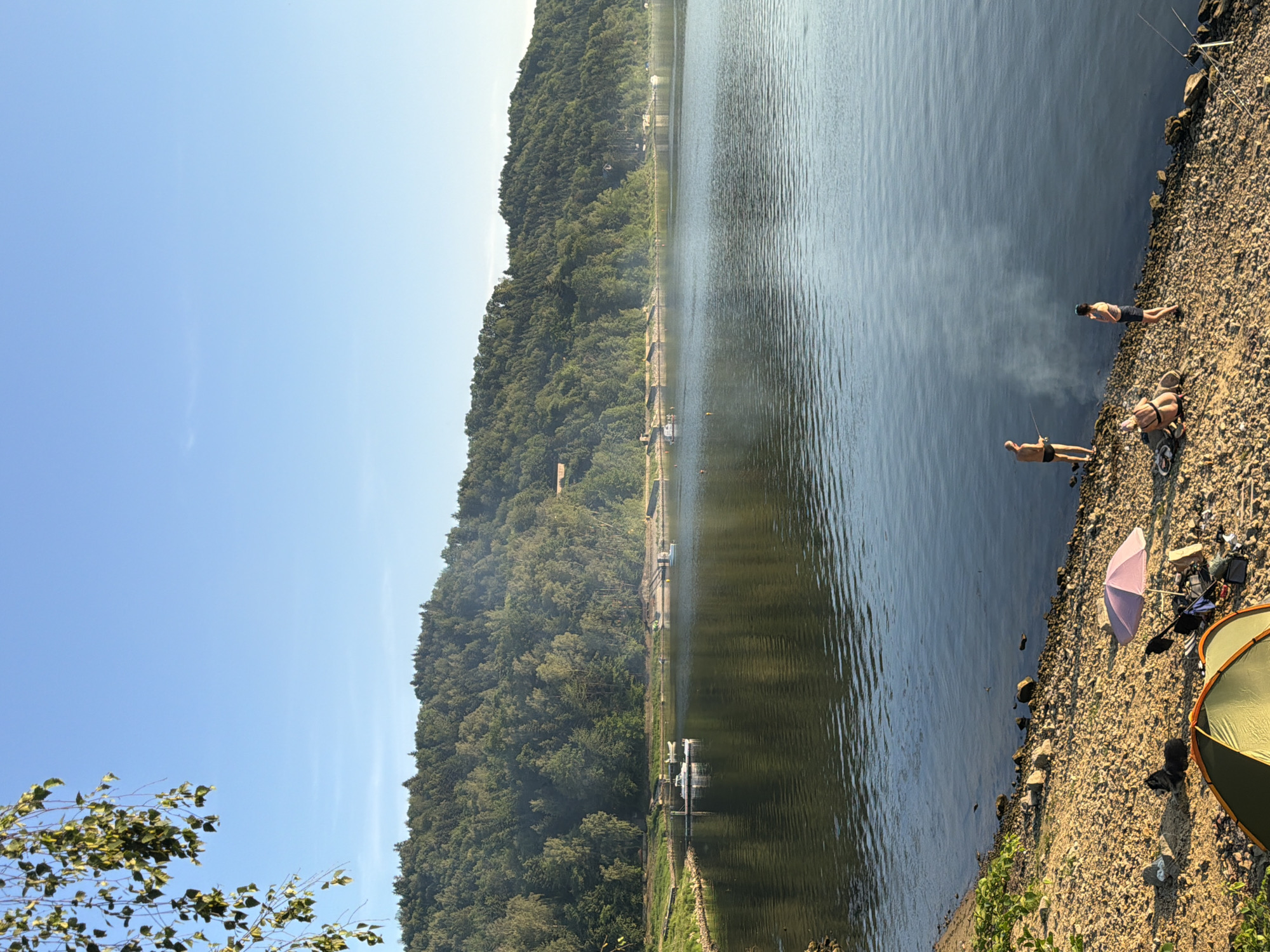 The image depicts a serene lakeside scene in Lipie, Poland. In the foreground, a rocky shore is dotted with camping equipment, including a green and orange tent, a pink umbrella, and various bags. Two individuals are visible near the water's edge: one person, wearing black shorts, stands and appears to be fishing, while another person in a striped top and dark shorts stands nearby. Further along the shore, another individual is seated, possibly preparing for fishing. A faint plume of smoke rises from the water between the two standing individuals. The lake itself is calm, reflecting the clear blue sky and the lush, tree-covered hillside that forms the backdrop. Several small boats are anchored in the distance along the shoreline. The overall atmosphere is one of tranquility and outdoor recreation.