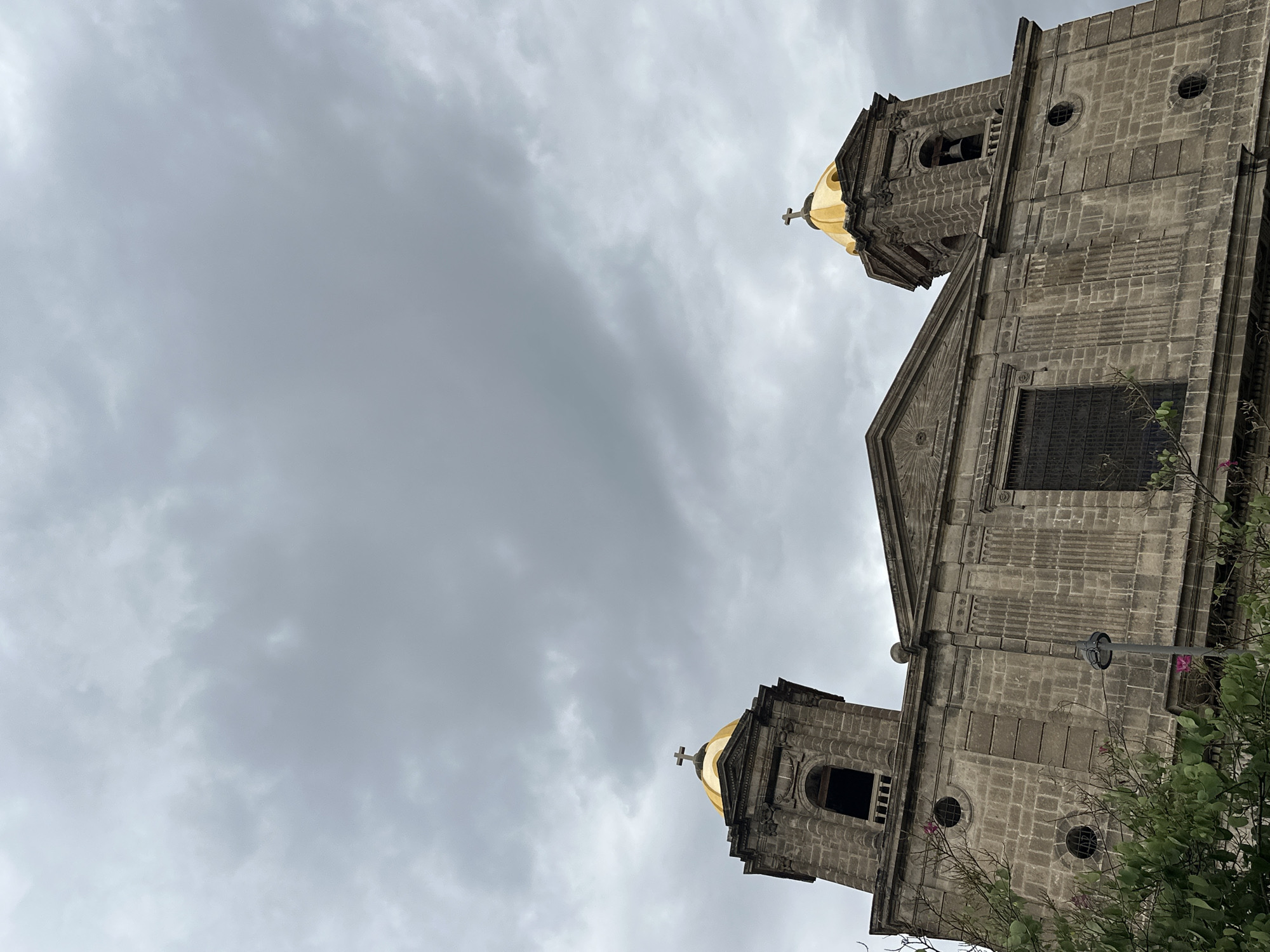 The image depicts the Metropolitan Cathedral in Mexico City, Mexico, viewed from a low angle looking upwards. The lower portion of the grey stone cathedral, including the two bell towers with golden domes and crosses on top, occupies the right side of the frame. The facade of the cathedral is visible, including a large window. Foliage is present in the lower right corner. The left side of the frame is dominated by a cloudy, overcast sky.