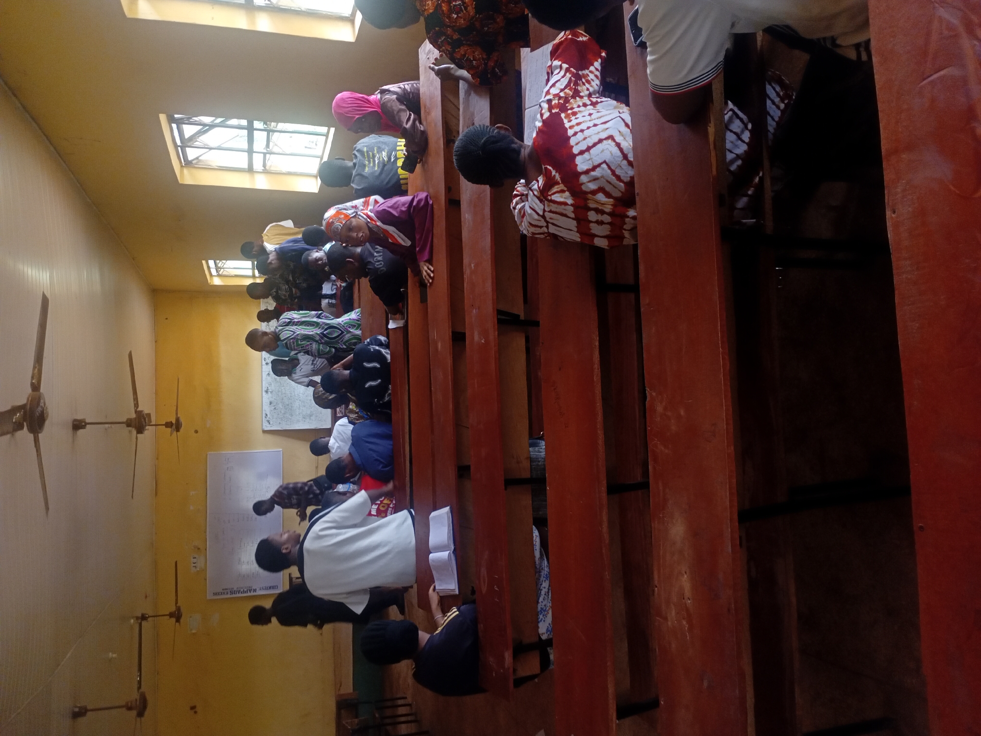 A classroom setting. Students are seated in rows of wooden desks. Some students are reading books. There are fans attached to the ceiling, and whiteboards on the wall. The location is Unknown, Unknown.