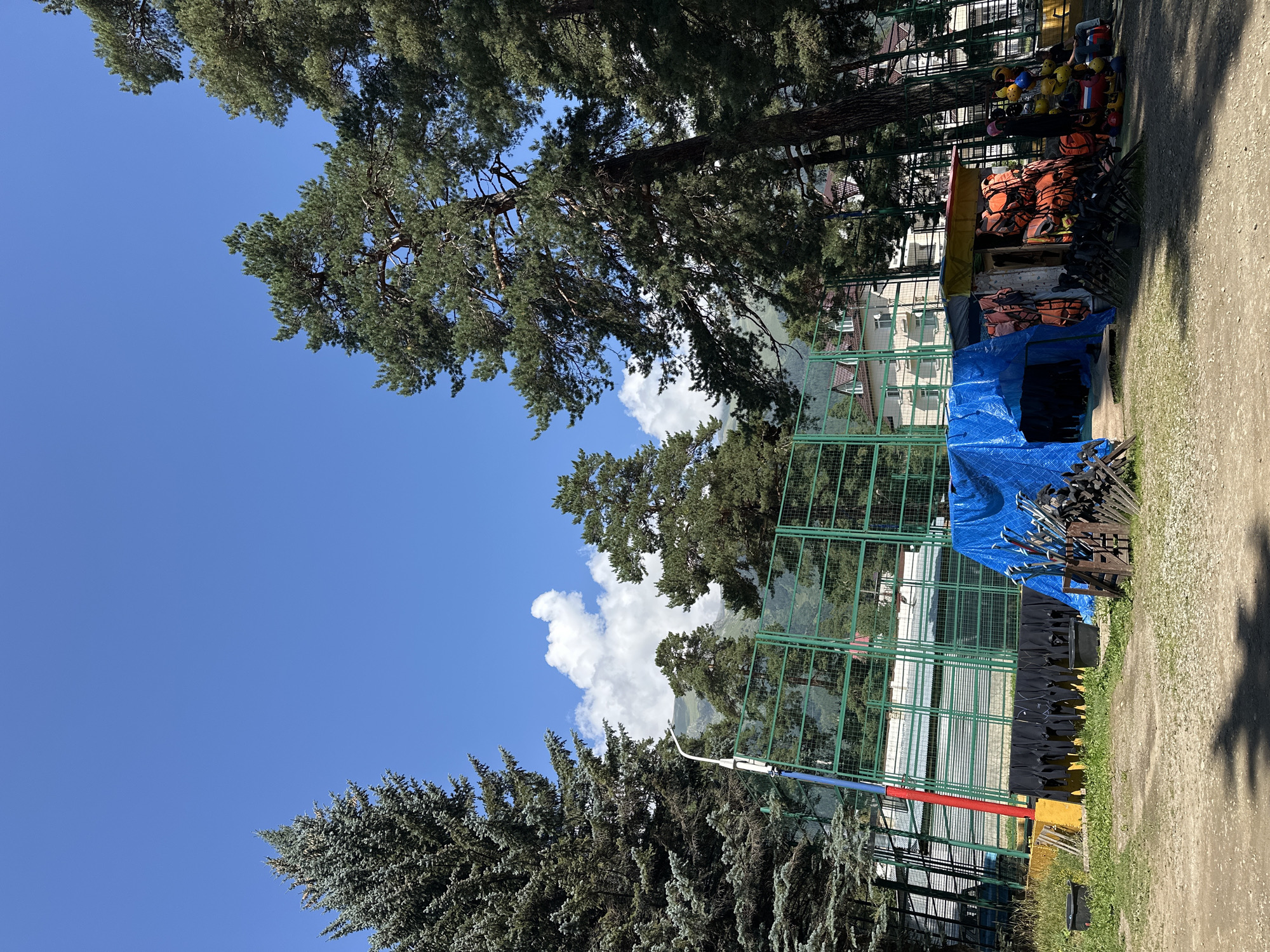 The image depicts an outdoor sports complex in Arkhyz, Russia. In the foreground, there is a gravelly area with grass patches. To the left, several black wetsuits are hung on a rack. In the center, a blue tarp covers some equipment. To the right, a small wooden structure is filled with orange life vests and colorful helmets. Behind this, a tall pine tree stands prominently. In the background, a green-fenced sports court is visible, with buildings and mountains beyond. The sky is bright blue with scattered white clouds.