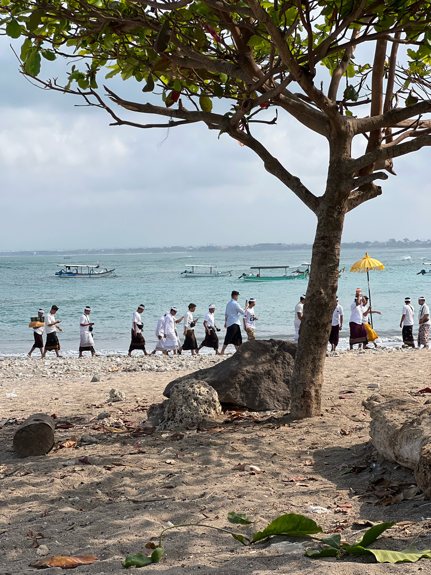 In Kuta, Indonesia, a group of men are walking along a sandy beach, with the ocean in the background. Most of the men are wearing white shirts and sarongs, and some have white headbands. One man in the middle is carrying a yellow umbrella over another man who is holding a tray above his head. Several boats are visible on the turquoise water. The beach is covered in sand, pebbles, and some driftwood. A large tree with green leaves partially obscures the scene in the foreground. The sky is overcast.