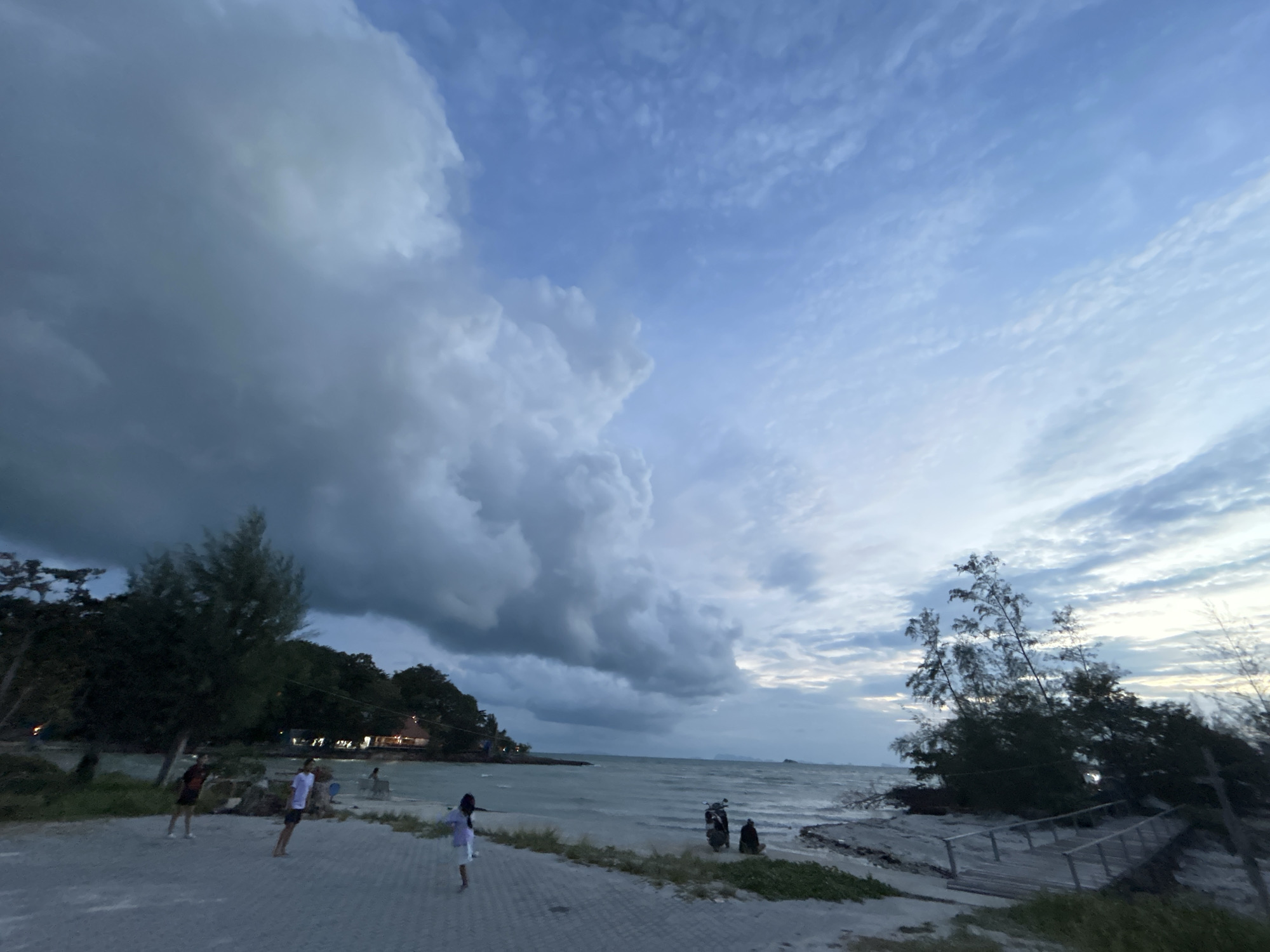 The image depicts a serene beach scene at dusk in Ko Pha-ngan District, Thailand. The sky is dominated by dramatic clouds, with a large, dark cloud formation on the left contrasting with lighter, wispy clouds on the right, suggesting an approaching storm or the aftermath of one. The sea is calm, with gentle waves reaching the shore.
On the paved walkway bordering the beach, several individuals are visible. Three people are standing and appear to be engaged in conversation or observing the surroundings. One person is further down the path on the right, facing away from the camera. In the background, along the coastline, there are trees and buildings, hinting at a developed area or a resort.
Near the water's edge, a scooter is parked, with two people seated nearby. The beach itself consists of sand and some sparse vegetation. A wooden walkway or pier extends from the beach into the sandier area on the right side of the frame, partially obscured by trees. The overall lighting indicates that it is either early morning or late evening, with the sky displaying a mix of blues and grays.