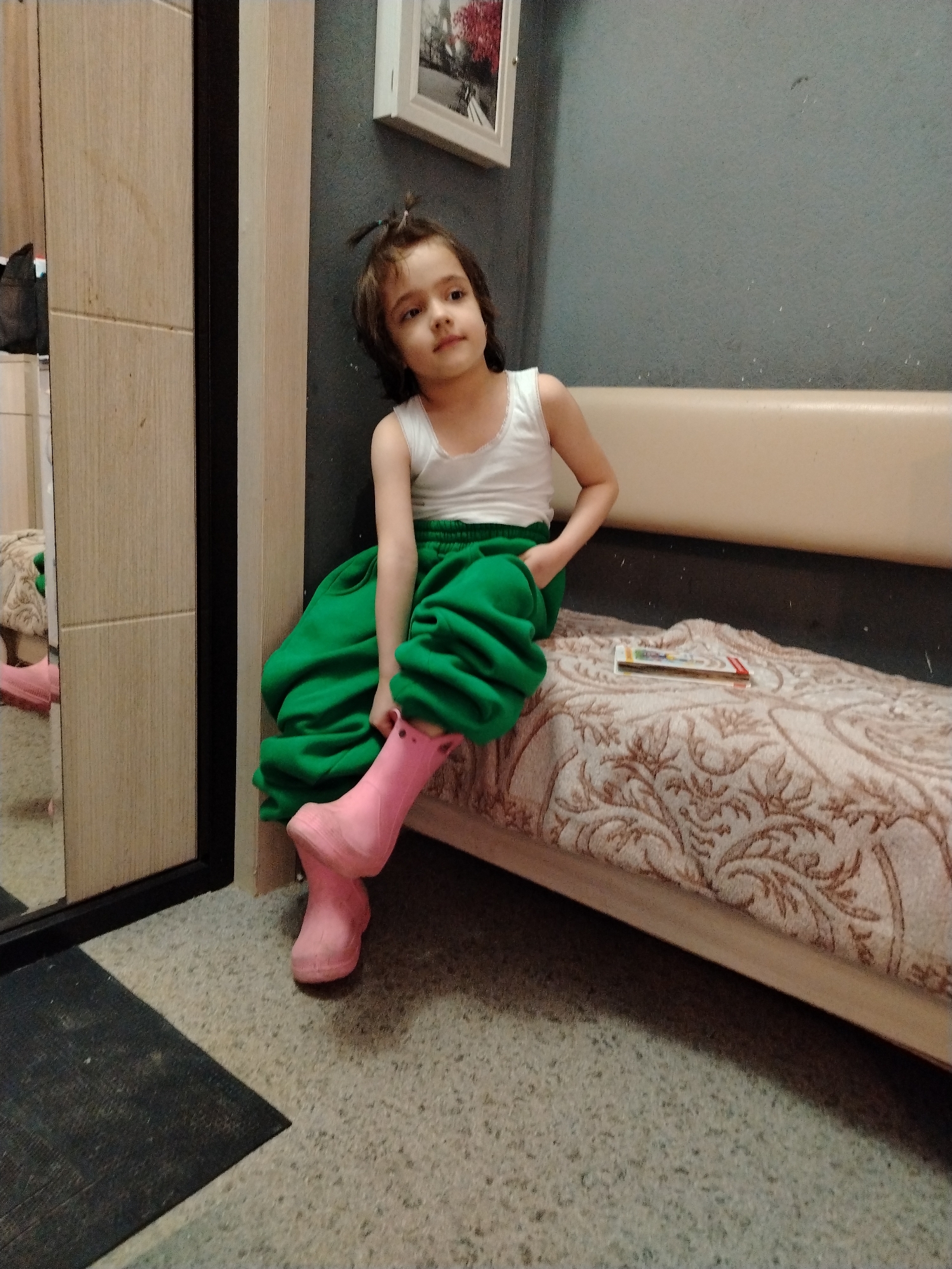 A young child with light skin, dark hair, and brown eyes sits on a bed or couch. She wears a white tank top, green baggy pants, and pink rubber boots. The child is looking to her left. A pack of crayons or colored pencils rests on the bed near her. A cabinet or closet with a mirror is to the left of the child. The room has a gray wall with a framed picture hanging on it. A dark mat or rug lies on the floor. The photo was taken indoors. I'm unable to verify the location in the city of Noviy, Russia, without additional information.