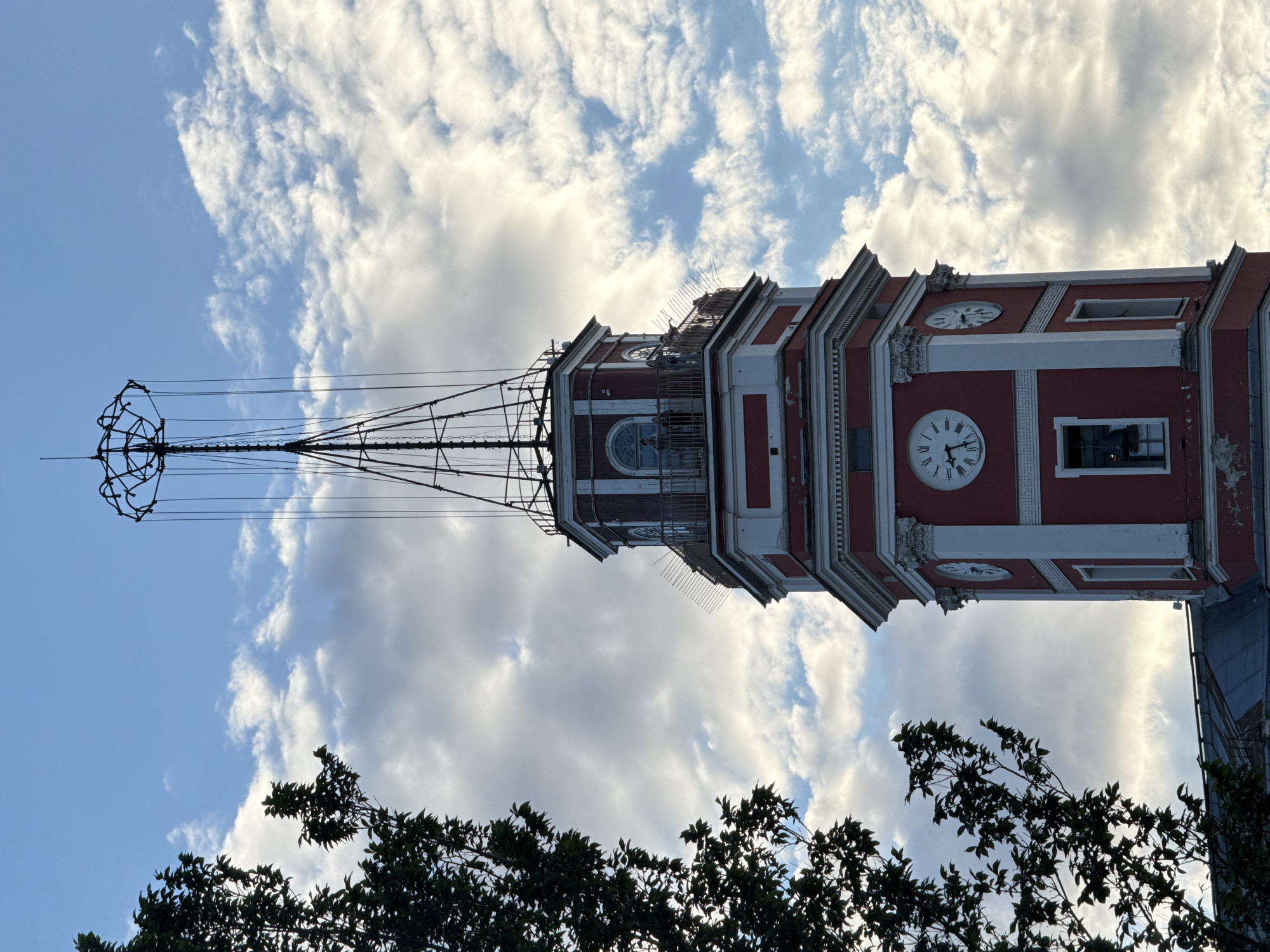 The image shows a close-up, low-angle view of the red and white tower of the Old Saint Petersburg Stock Exchange building, with a tree on the left. The tower has several clocks on its octagonal faces and a unique antenna structure at the top. Several people are visible standing on a balcony near the top of the tower. The sky is blue with scattered white clouds, and the sun is casting a warm light on the building. The tree on the left is lush and green. The building's architectural style is neoclassical, typical of St. Petersburg.