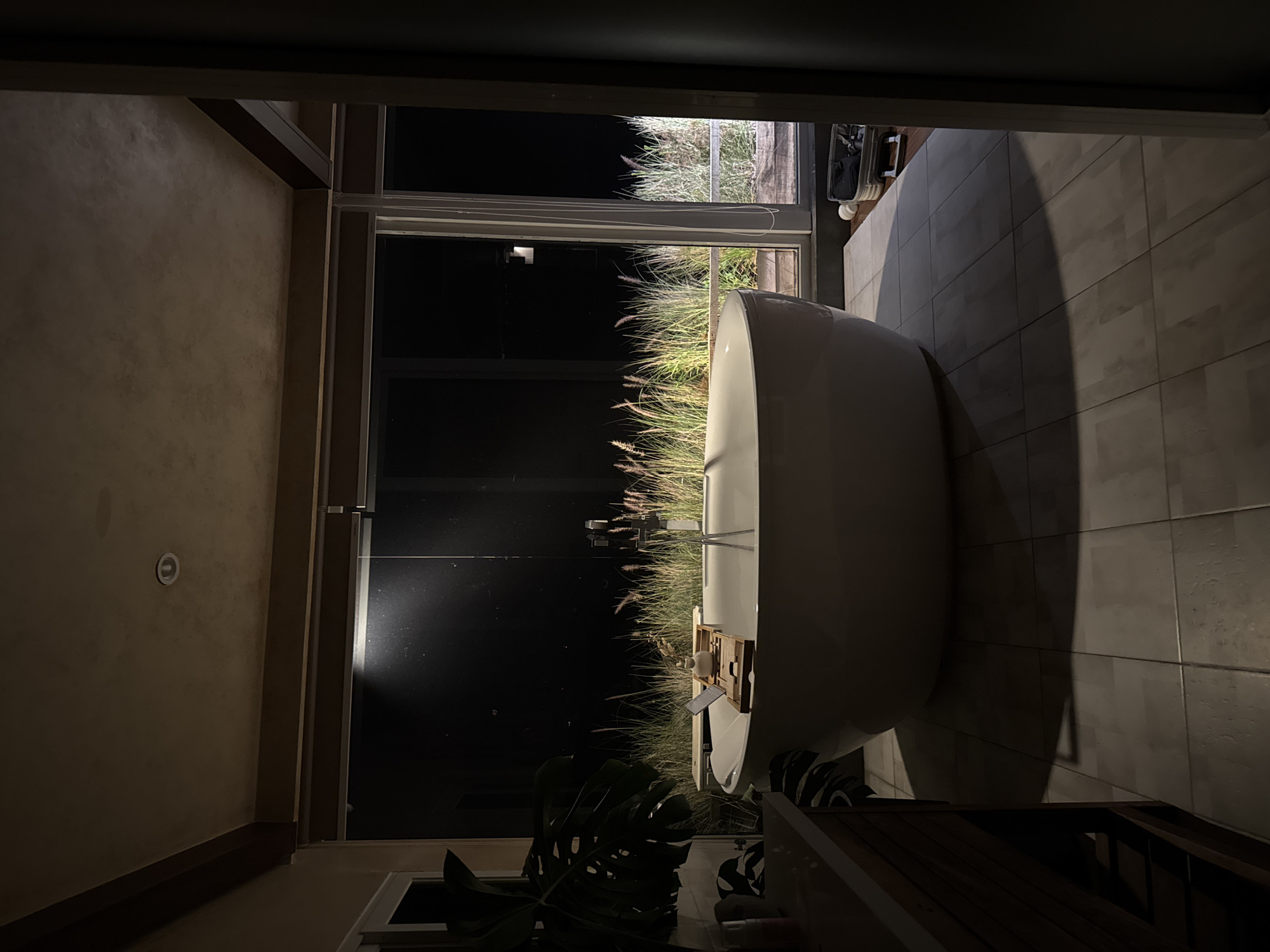 A freestanding white bathtub with a wooden bath tray sits in a dimly lit bathroom. The tray holds a small white vase and a small book. Behind the bathtub, large windows offer a view of tall, dry grasses against a dark night sky. To the left of the bathtub, a large, dark green leafy plant is visible, and to the right, a suitcase rests on the tiled floor. The floor is tiled with square, light gray tiles. The scene is located in a bathroom in Da Lat, Vietnam.