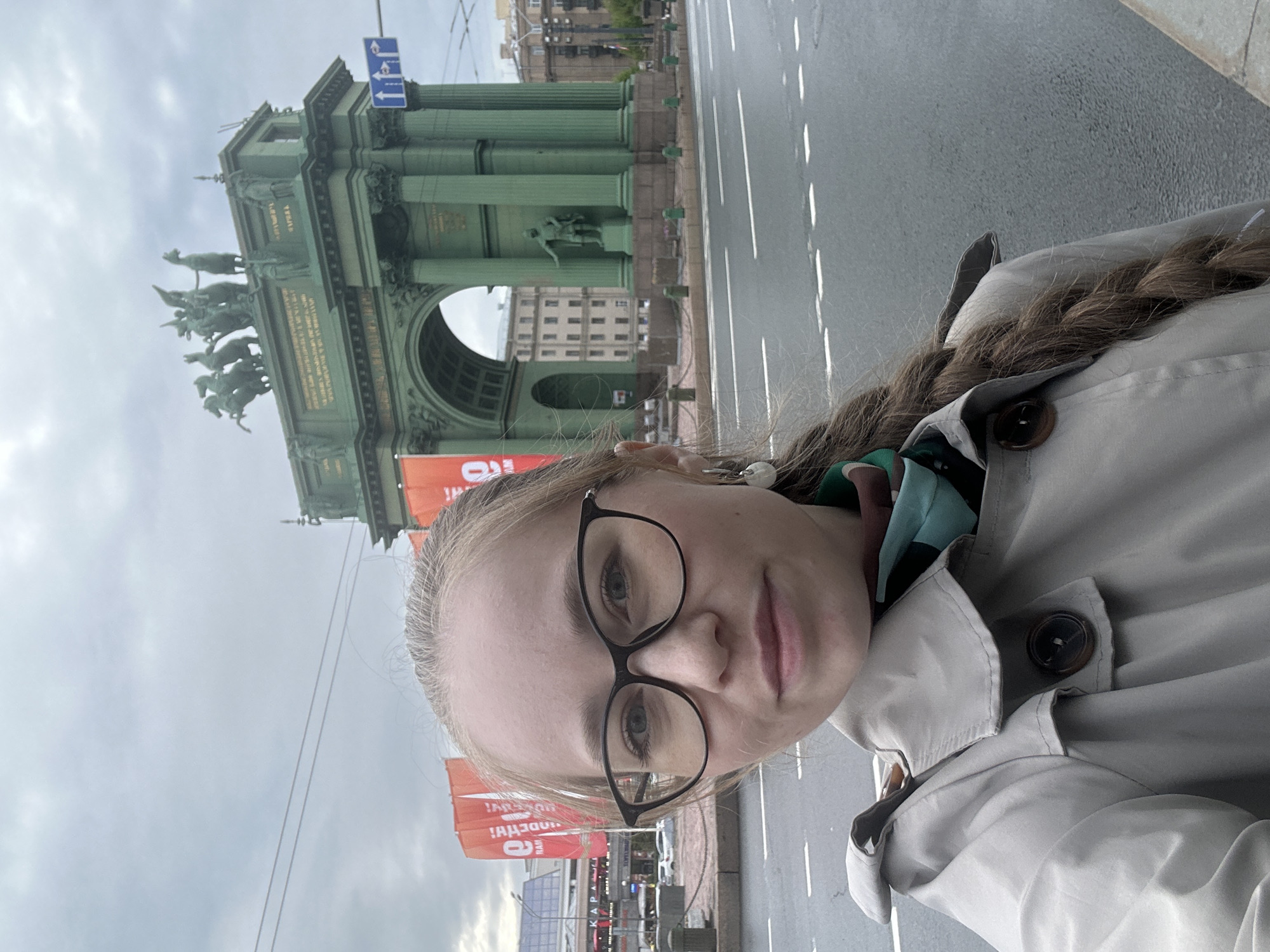 A woman with light-colored hair, wearing glasses and a beige trench coat, is taking a selfie on a street in Saint Petersburg, Russia. In the background is the Narva Triumphal Arch, a historical monument featuring green columns and sculptures. The street appears wet, with visible lane markings. Banners and signage are present in the distance.