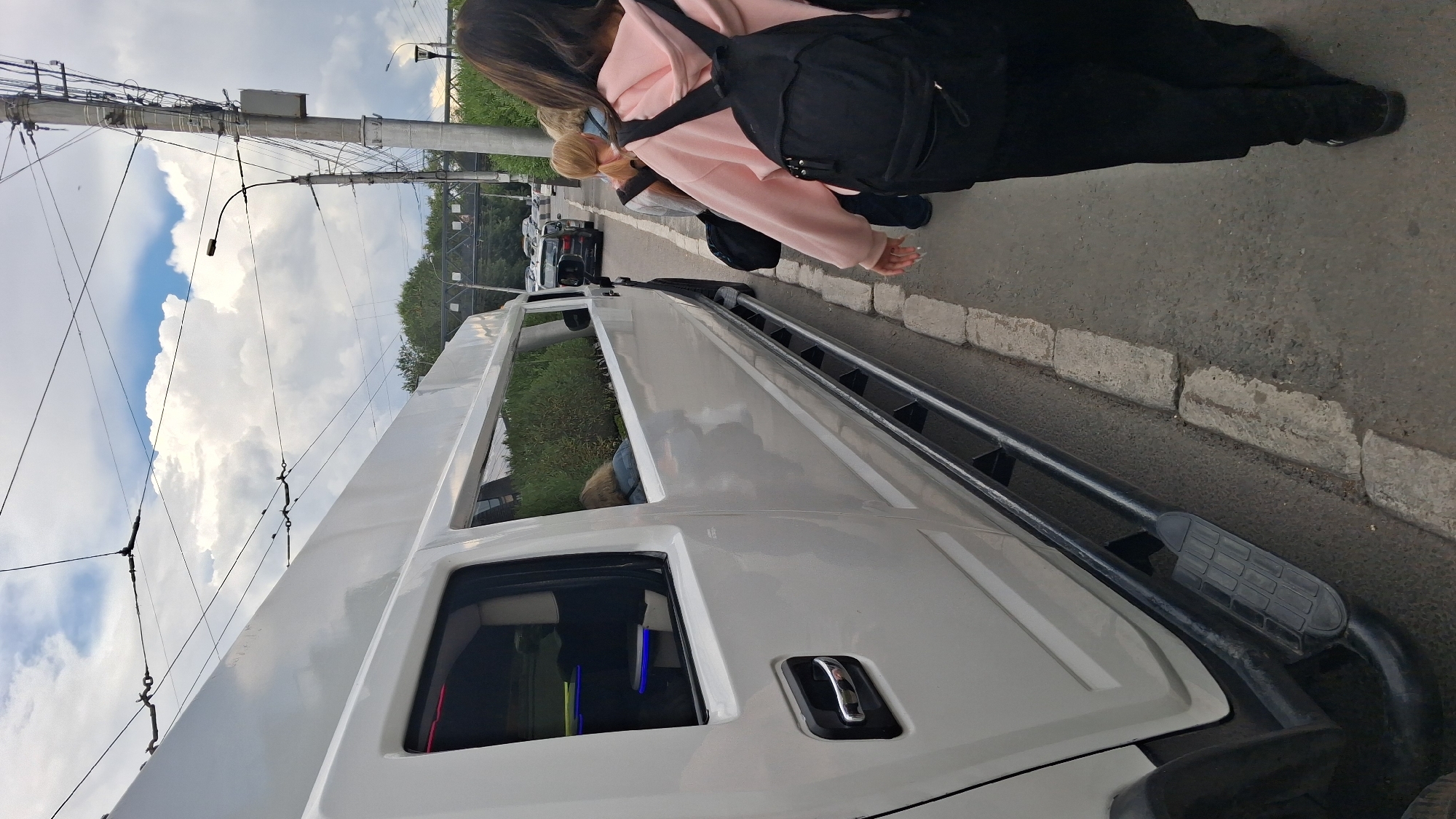 A white stretch limousine is parked on the right side of the road, adjacent to a sidewalk. Several people are walking away from the limousine on the sidewalk. The road appears to be a city street with overhead electrical wires and streetlights visible. In the background, there are more vehicles on the road and some trees. The sky is cloudy. The scene is likely in Novosibirsk, Russia, given the presence of overhead electrical wires commonly used for public transport in Russian cities.