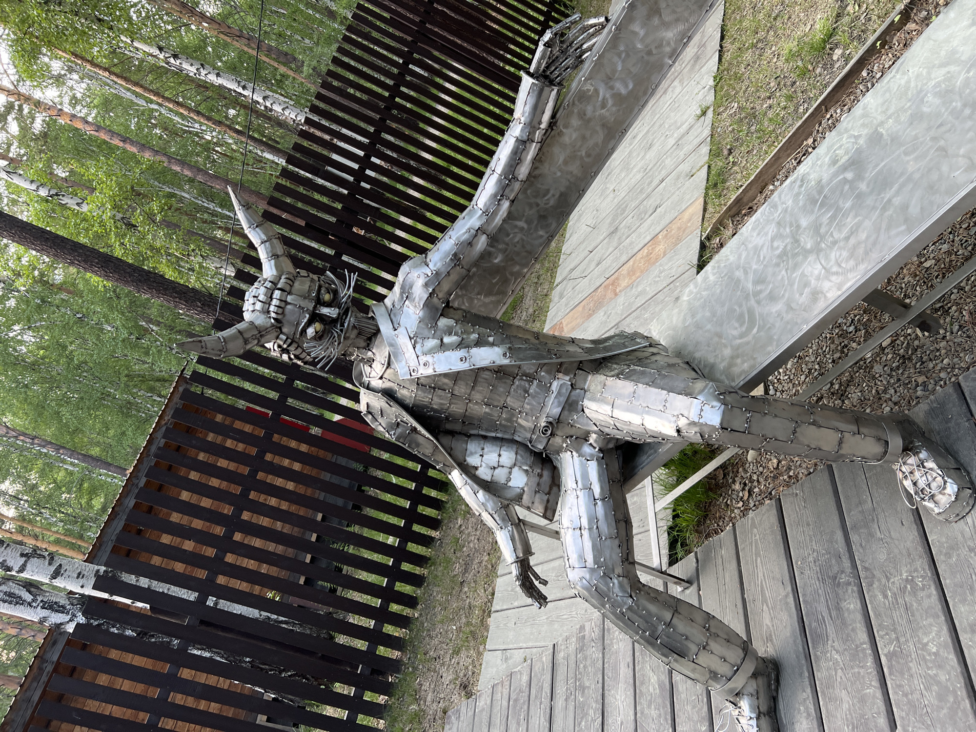 A full shot shows a metal sculpture of a seated figure with a horned helmet. The sculpture is made of numerous small, riveted metal plates, giving it a segmented appearance. The figure is wearing a suit jacket and trousers, and its arms are outstretched. The sculpture is sitting on a metal bench in an outdoor setting. The background consists of a wooden fence, a forest with birch trees, and a wooden deck. The location is the Taltsy Museum of Architecture and Ethnography in Tal'tsy, Russia.