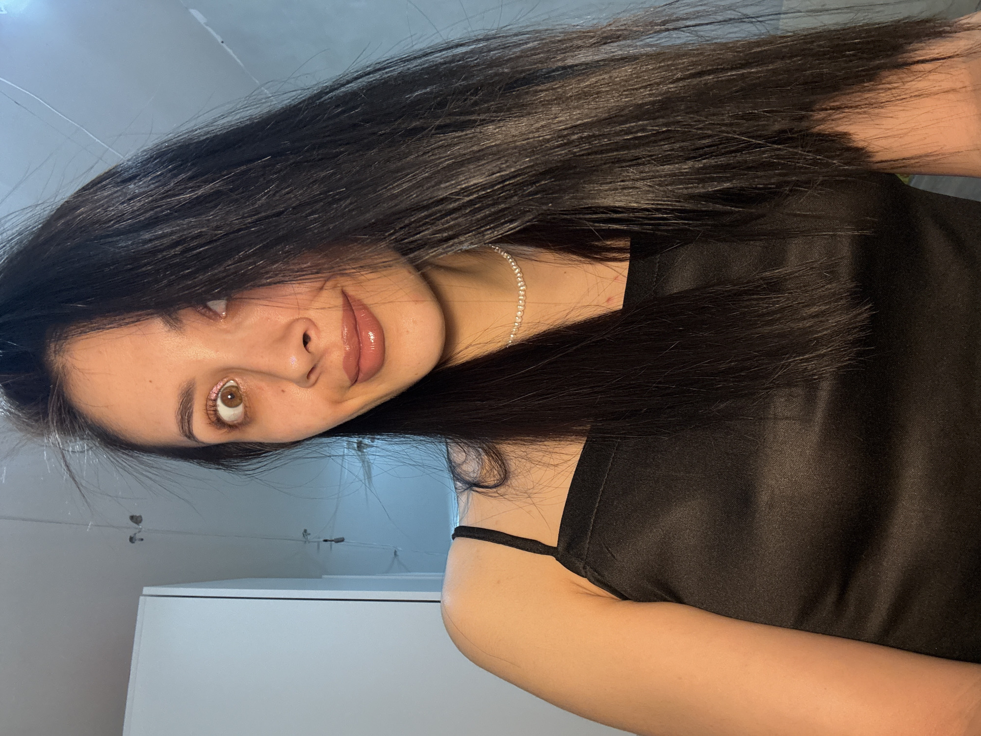 A young woman is captured in a close-up, casual portrait, likely a selfie, in Votkinsk, Russia. She has long, straight, dark hair that falls loosely around her, partially obscuring her face and extending across her shoulder. Her head is tilted, and she gazes directly at the viewer with light brown eyes, one eyebrow slightly raised, conveying a playful or confident expression. Her lips are full and enhanced with a glossy, light pink or nude lipstick, forming a subtle smile or smirk. She wears a black top or dress with thin straps, possibly made of satin or silk, given its sheen. A delicate, silver-toned choker necklace is visible around her neck. The background is minimalistic, featuring a light-colored wall or ceiling and a white piece of furniture, possibly a cabinet or dresser, in the lower left. Some thin wires or lines are also visible on the wall above her. The lighting is soft and ambient, suggesting an indoor setting without specific time-of-day cues. There is no visible text.
