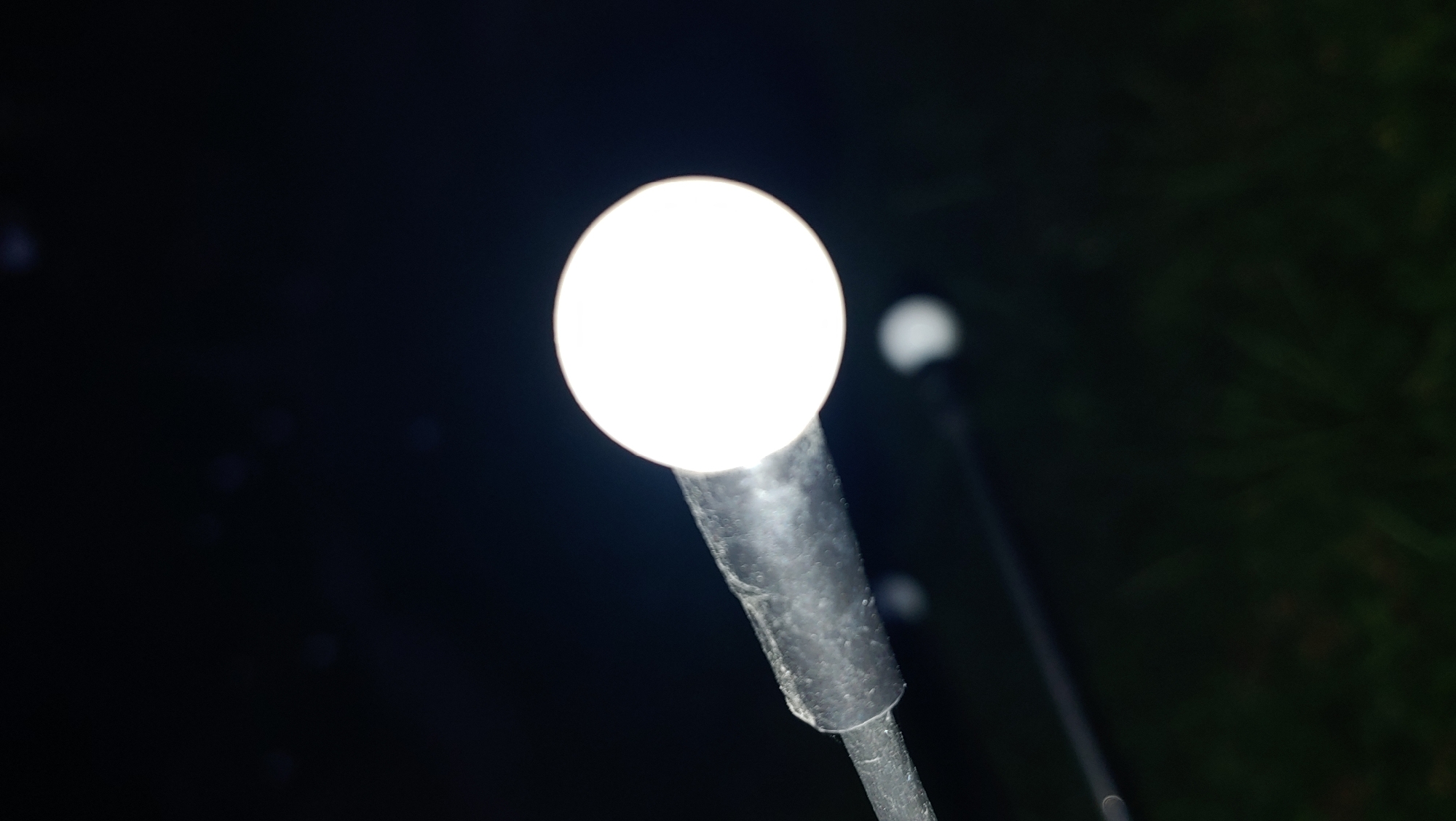 An out-of-focus, close-up shot of a bright, round LED light fixture is captured against a dark, blurry background. The light source appears to be attached to a dark, textured pole, which extends diagonally from the left side of the frame. The background consists of indistinct shapes and colors, suggesting a dark environment, possibly outdoors with some distant lights or reflections. The lower portion of the image shows a blurred green and dark texture, likely indicating grass or foliage. There is a second, fainter, out-of-focus light source visible in the background, to the right of the main light. There is no discernible information about the city of Mukachevo, Ukraine, or any people, actions, or interactions within this image.