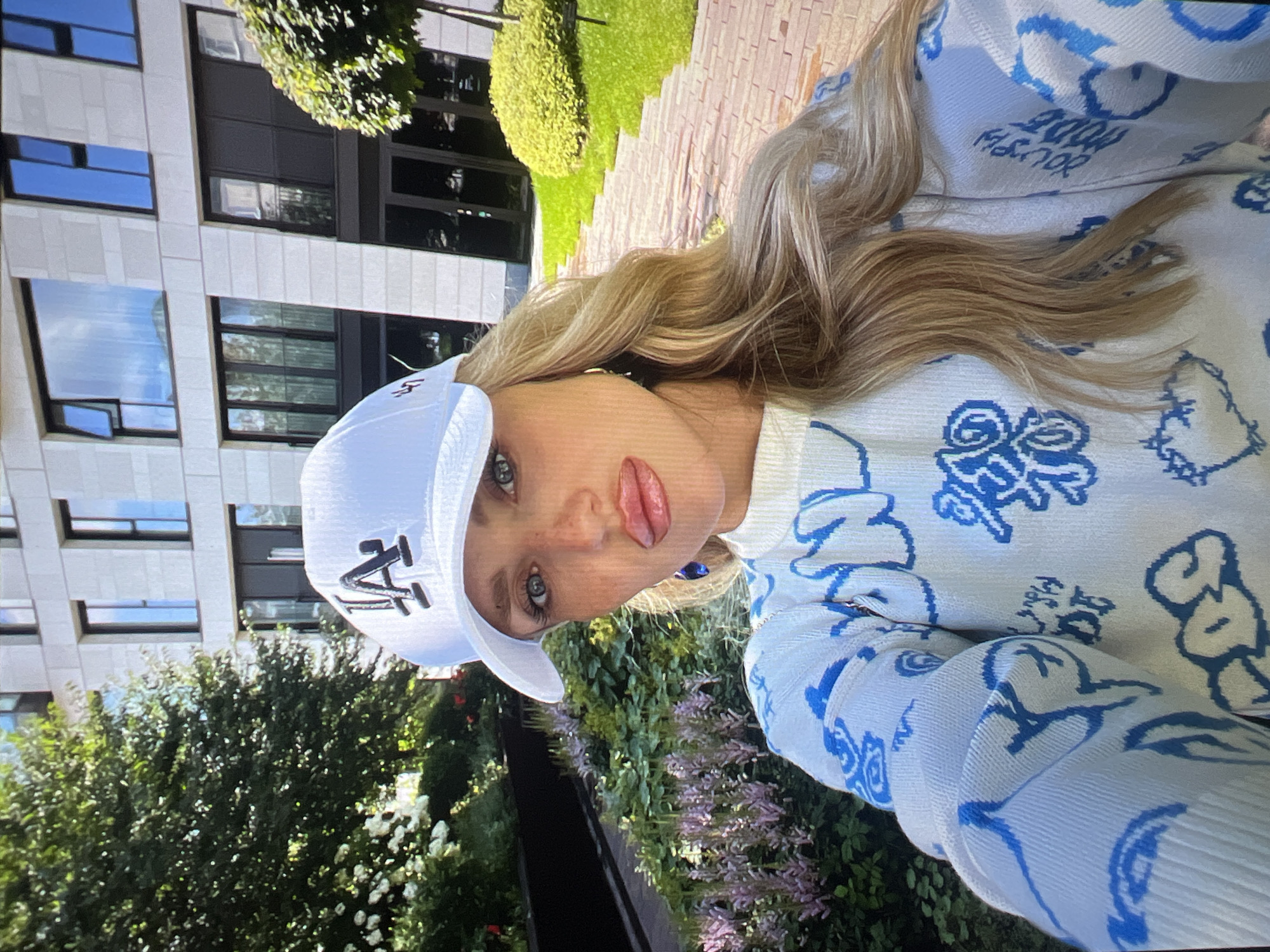 A blonde woman wearing a white baseball cap with a black "LA" logo and a white sweater with blue graffiti-style designs is taking a selfie outdoors. She has full lips, light skin with freckles, and blue eyes. She is standing in front of a modern, light-colored building with large windows, surrounded by lush green trees and a flowerbed with purple flowers. A brick pathway is visible to her right. The scene appears to be a sunny day. The city is Unknown, Russia.
