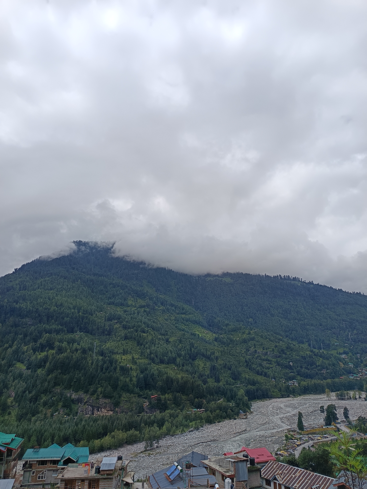 The image captures a wide view of a mountainous landscape under a cloudy sky, likely in the vicinity of Vashisht, India. In the foreground, a cluster of buildings with predominantly green and red roofs are visible. These structures appear to be residential or small commercial establishments, with varying architectural styles. A rocky riverbed, likely fed by a stream or river, cuts through the middle ground, with a road winding alongside it. The background is dominated by a densely forested mountain slope, with a significant portion of the mountain's peak obscured by low-hanging clouds. The overall atmosphere is overcast and humid, suggesting a possibly rainy or post-rainy season environment. No people or active events are depicted in the scene.