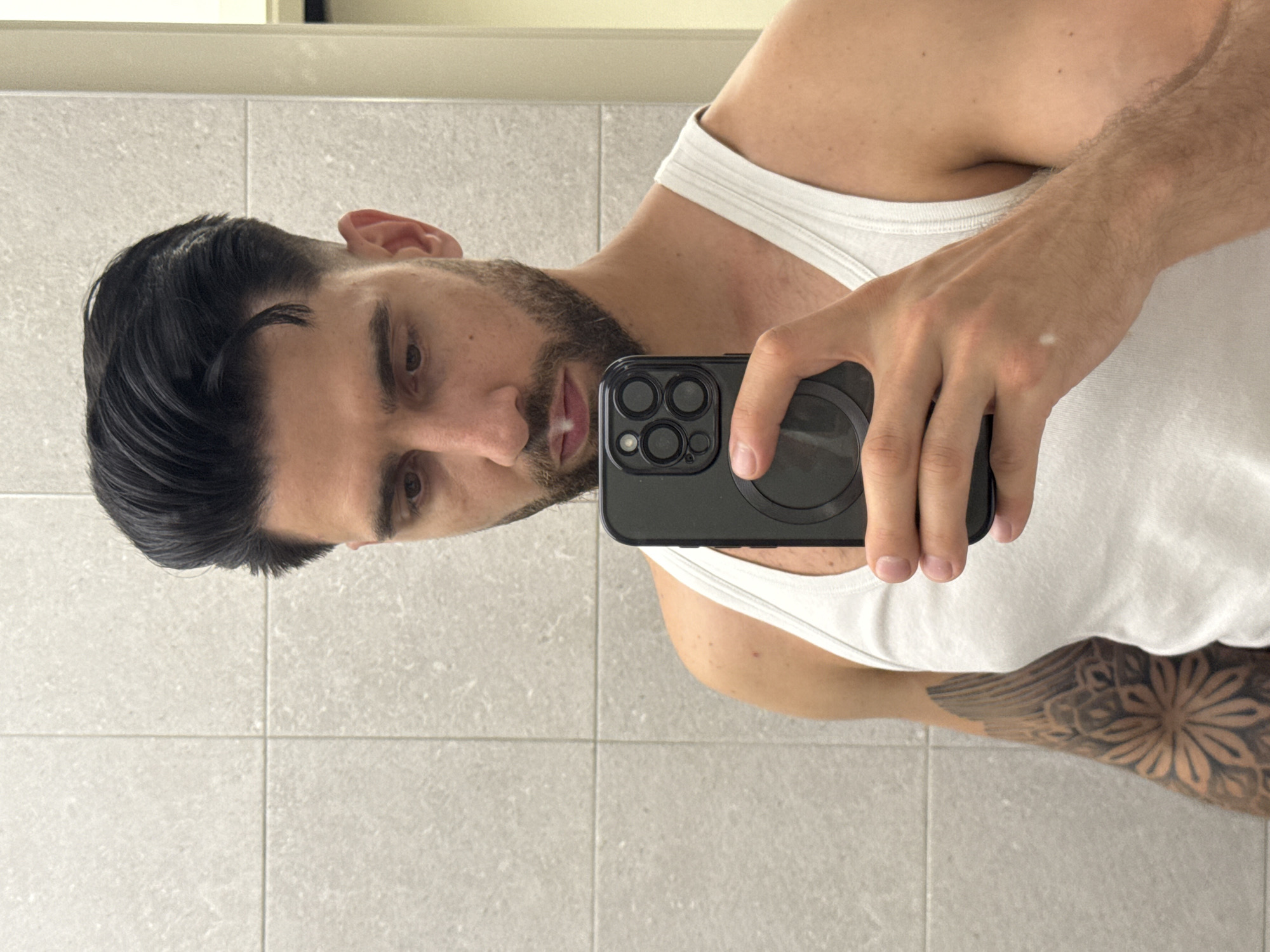A man is taking a selfie in a bathroom. He is wearing a white tank top and holding a black smartphone. He has dark hair, a beard, and a tattoo on his arm. The bathroom has light-colored tiled walls. There is no information available about the location in the city of Sofia, Bulgaria.