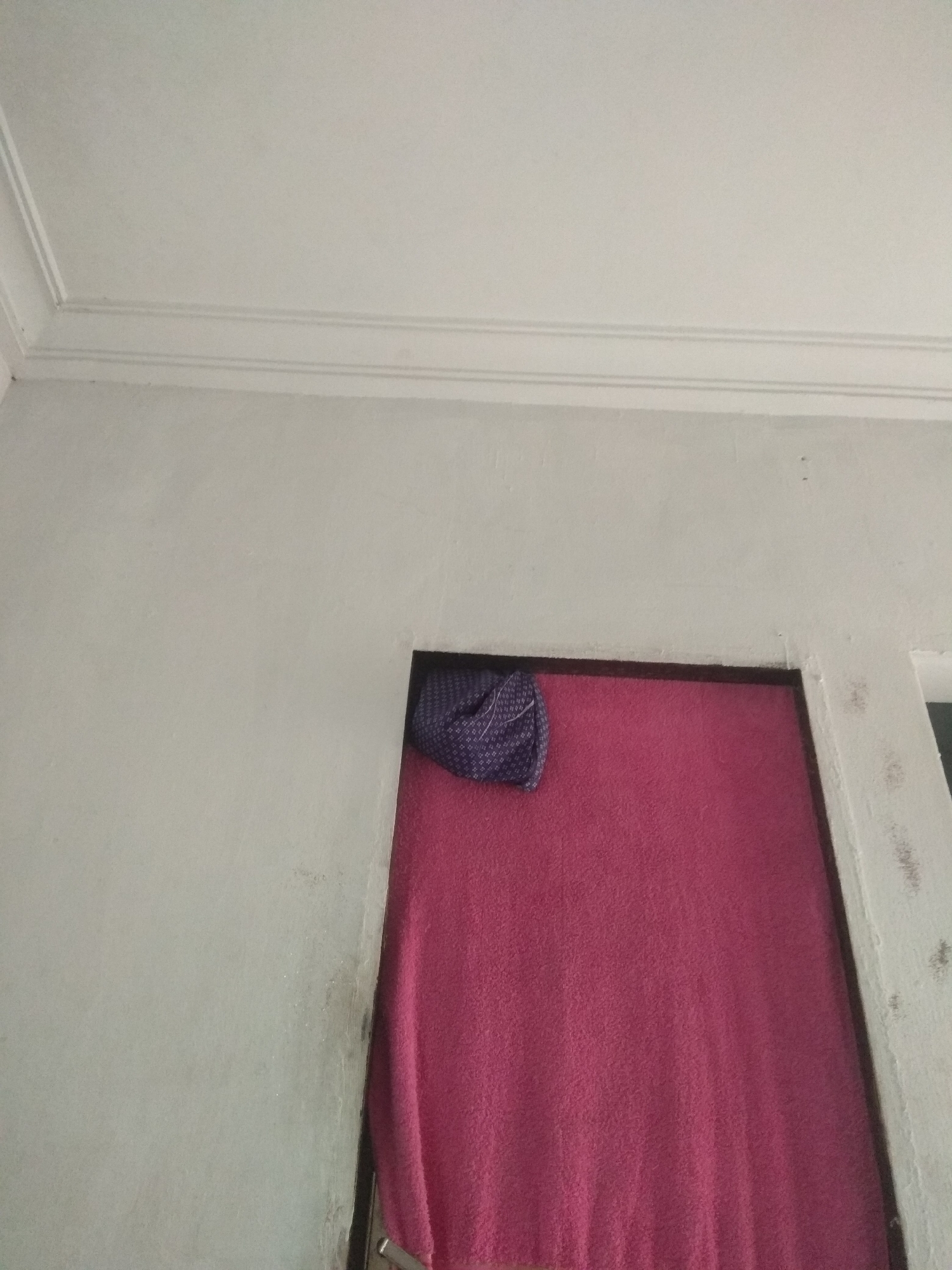 The image depicts an interior view of a room with a white ceiling featuring decorative molding. A wall, painted a light gray, shows some signs of wear and discoloration, particularly near a doorway. The doorway is partially obscured by a vibrant pink towel or fabric draped within its frame. A small portion of a purple patterned cloth is visible near the top left of the pink fabric, peeking out from behind it. The overall scene suggests an ordinary domestic setting, possibly within a residential building in Jalingo, Nigeria, though no specific landmarks or external environmental cues confirm the location. The focus is on the textures and colors of the wall, ceiling, and the draped fabrics.