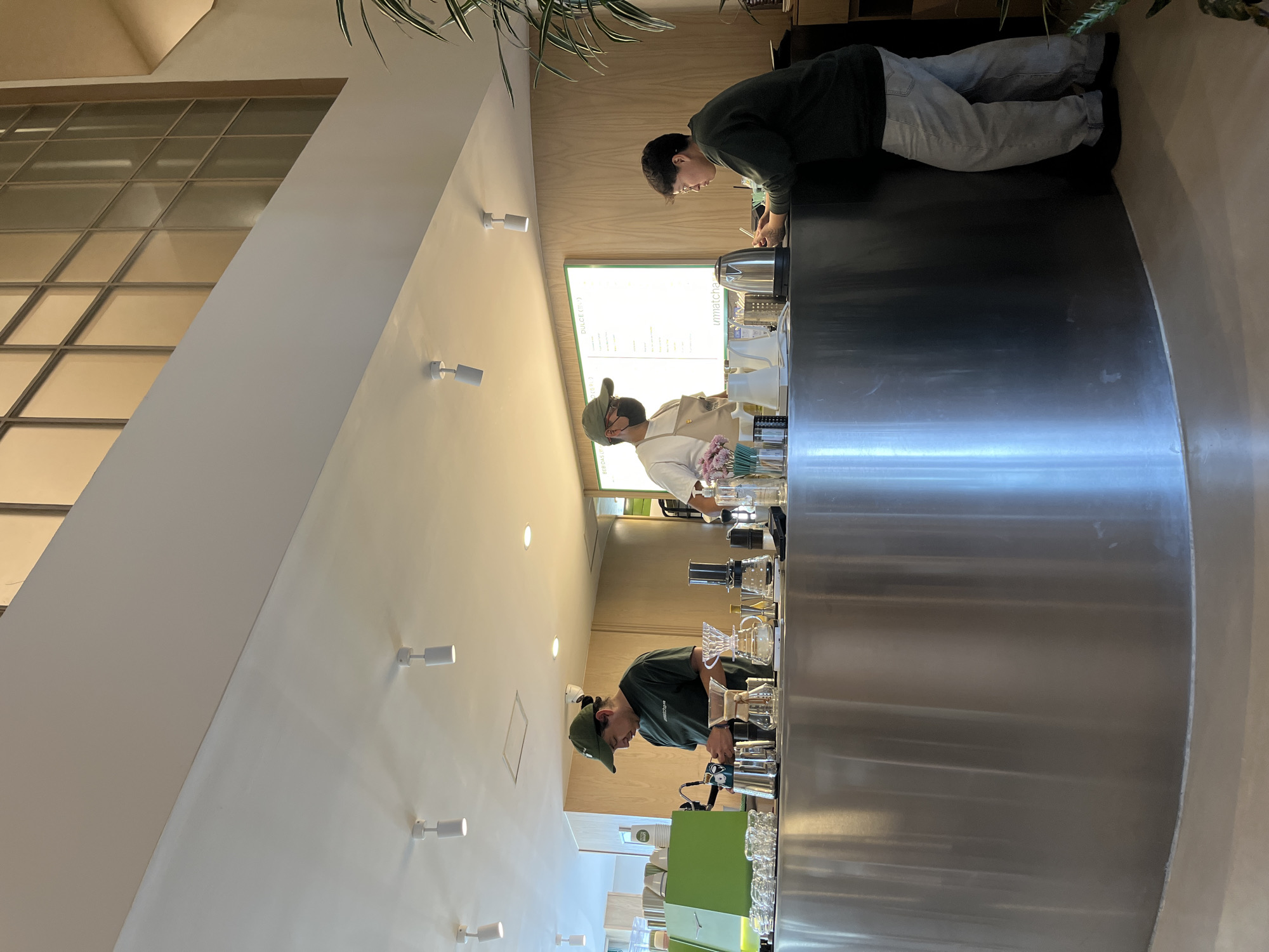 Inside an unidentified business location in an unknown city, three individuals are positioned behind a curved, stainless-steel countertop. The individual closest to the camera is wearing a green cap and a dark green shirt, appearing to be engaged in some activity at the countertop. Further back, another person is wearing a light-colored outfit and a cap. The third individual, wearing a dark top and light pants, is standing at the end of the countertop. Various objects are arranged on the counter, including glassware, a kettle, and other unidentified equipment. The background features a wall with small lights, a menu board, and a window at the top left of the frame. The lighting is bright. A plant hangs from the ceiling at the top of the frame.