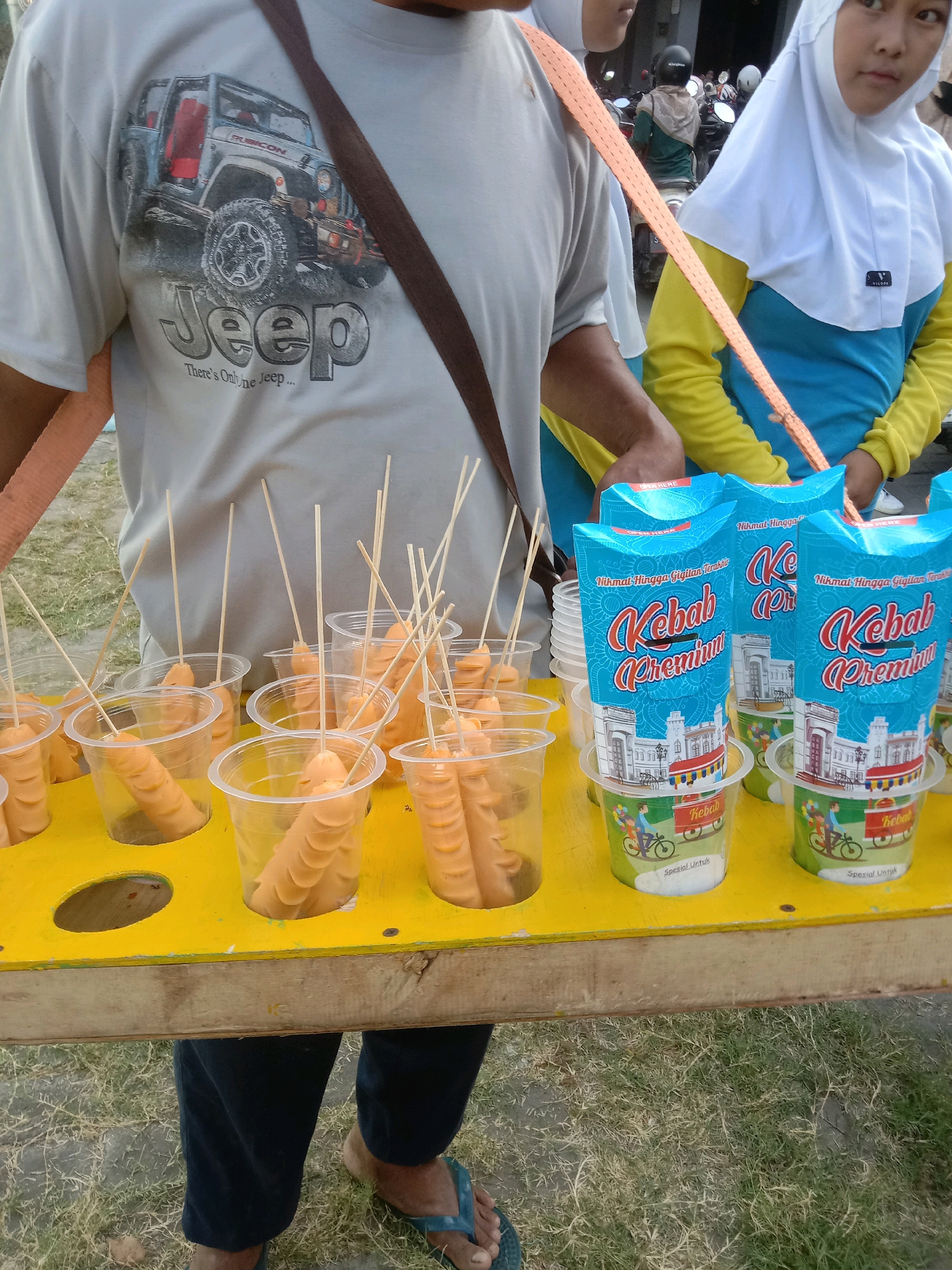 In Sumberrejo, Indonesia, a street vendor displays a variety of hot dogs and kebabs. The vendor, wearing a grey t-shirt featuring a Jeep design, stands behind a yellow wooden cart laden with food items. Several plastic cups filled with hot dogs on skewers are arranged in the cart, alongside blue packaging for "Kebab Premium." Two young women, one in a yellow and blue hijab, are positioned to the right of the vendor, potentially customers or assistants. The background reveals a street scene with parked motorcycles and other pedestrians, suggesting a busy urban environment. The ground is covered in dry grass and dirt, indicative of outdoor market conditions.