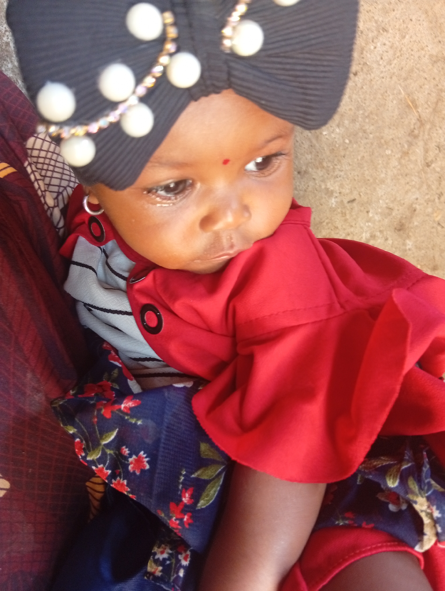 A close-up, eye-level shot shows a young child's face and upper body. The child, likely a baby, wears a black turban adorned with white beads and a beaded headband. A red bindi is visible on the child's forehead. The child has large, dark eyes with visible eyelashes and is looking to the right. The child's left ear, adorned with a silver hoop earring, is visible. The child is wearing a red top with decorative black circles and a striped fabric underneath. A portion of a blue fabric with a red and white floral pattern is also visible. The background is a textured, light-colored surface, possibly concrete or stone.