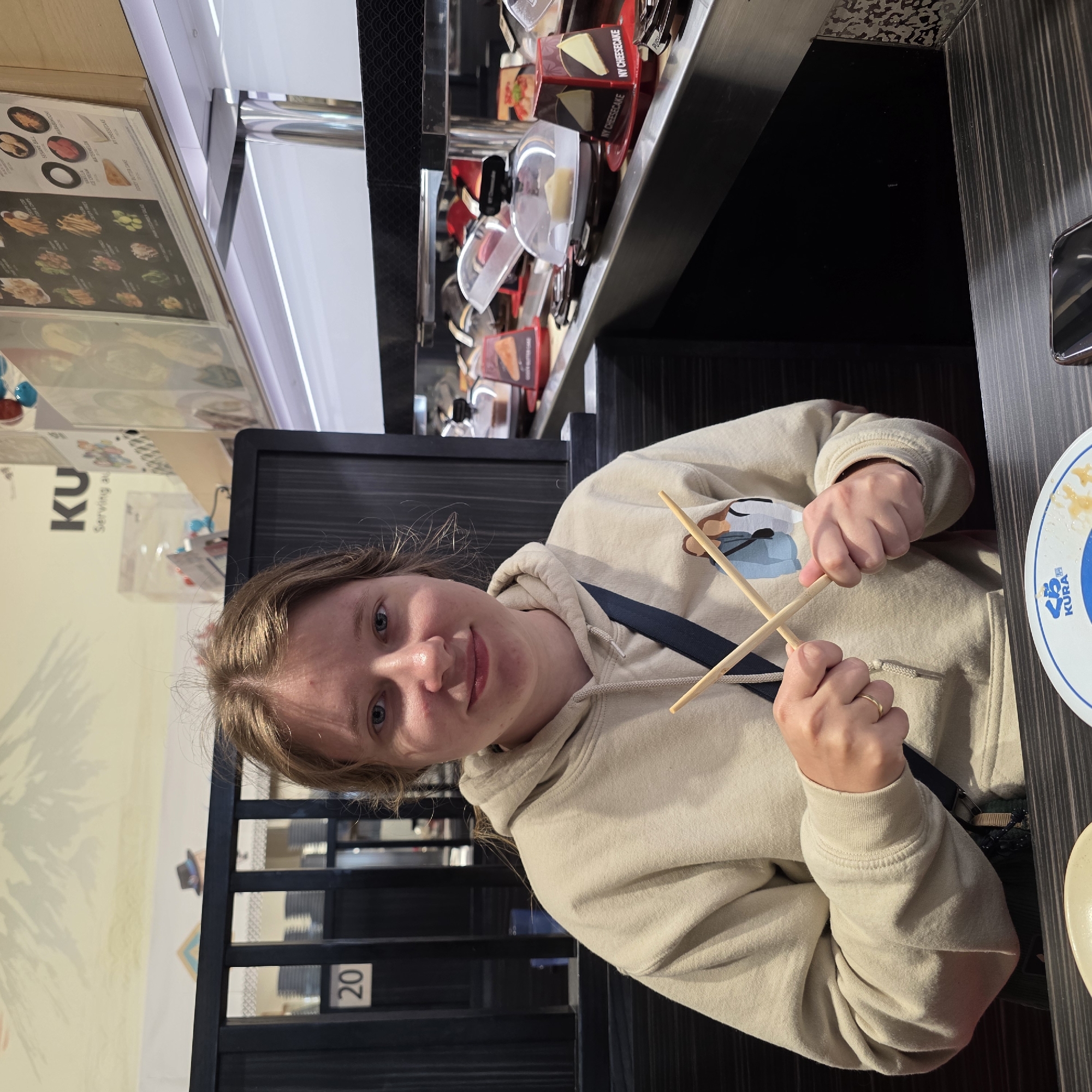 A young Caucasian female with shoulder-length blonde hair and blue eyes is seated in a booth at a Kura Sushi restaurant in Beaverton, Oregon, USA. She is wearing a light beige hooded sweatshirt with a printed graphic of two figures on the front. Her hands are clasped together, holding a pair of wooden chopsticks. She is smiling and looking directly at the camera. In the background, a conveyor belt system displays various food items in plastic containers, including slices of cheesecake. The restaurant features dark wood paneling and a mural of a mountain scene on the wall behind the booth. The table in front of her has a white plate with blue lettering that reads "KURA".