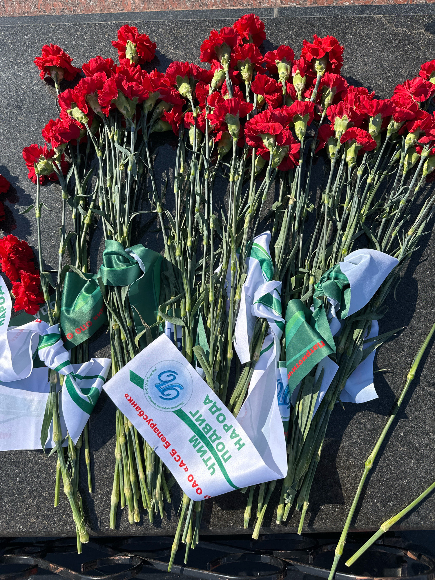 A bouquet of red carnations with green ribbons and white sashes with Cyrillic lettering is laid on a dark, polished stone surface, likely a monument or gravestone. The stone appears to be granite. The sashes bear inscriptions that read "ПОБЕДЫ НАРОДА" (Victory of the People) and "ОАО «АСБ Беларусбанк»" (JSC "ASB Belarusbank"). One sash also features a blue logo. The carnations are tightly packed, with their green stems visible. The lighting suggests it is daytime, with shadows cast on the stone. This scene is likely at the Brest Fortress, a significant historical site in Brest, Belarus, commemorating World War II.