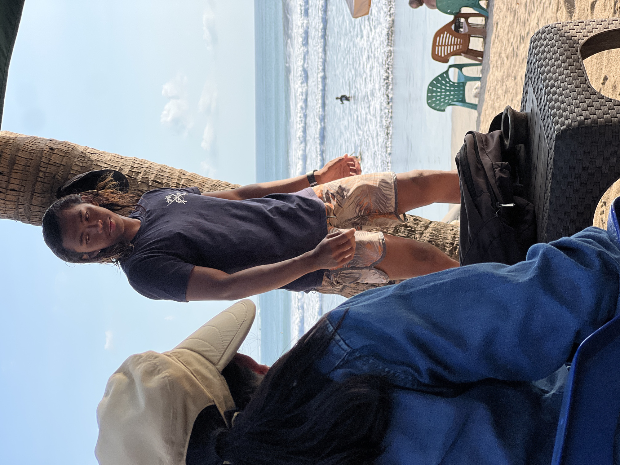 In Kuta, Indonesia, a young man with long, dark hair and a navy t-shirt stands leaning against a palm tree on a sandy beach. He wears patterned shorts and a watch on his left wrist. In the foreground, from the back, a person wearing a blue denim jacket and a light-colored hat is seated. A dark bag and a small dark bowl are on a woven plastic table in front of them. Beyond them, the ocean with gentle waves stretches to the horizon under a clear blue sky with scattered clouds. Several colorful plastic beach chairs are visible on the sand. A distant figure can be seen in the water.