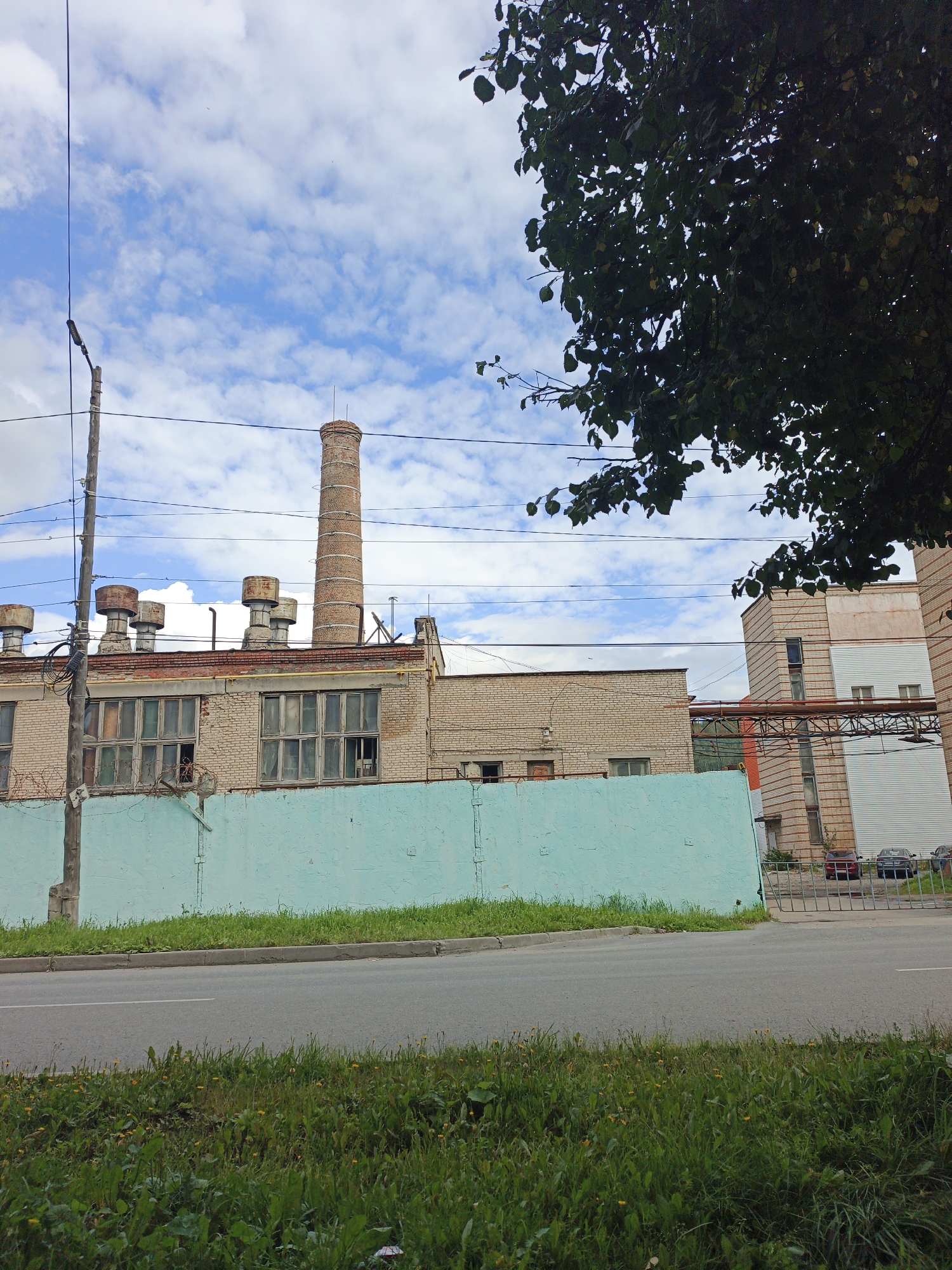 The image depicts an industrial building with a tall brick smokestack and several smaller ventilation units on its roof. The building is constructed of light-colored bricks and features numerous large windows, some of which appear broken or boarded up. A light blue, rough-textured concrete wall runs along the front of the property, separating it from a paved road. Beyond the wall, a grassy area with some wildflowers is visible. In the background, another industrial structure with a multi-colored facade and white corrugated metal siding can be seen, along with a metal gate and a few parked vehicles. Overhead, a network of power lines is strung between utility poles, and the branches of a leafy tree are visible in the upper right portion of the frame against a cloudy blue sky. The scene appears to be in an urban industrial area, and the overall impression is one of disuse or age.