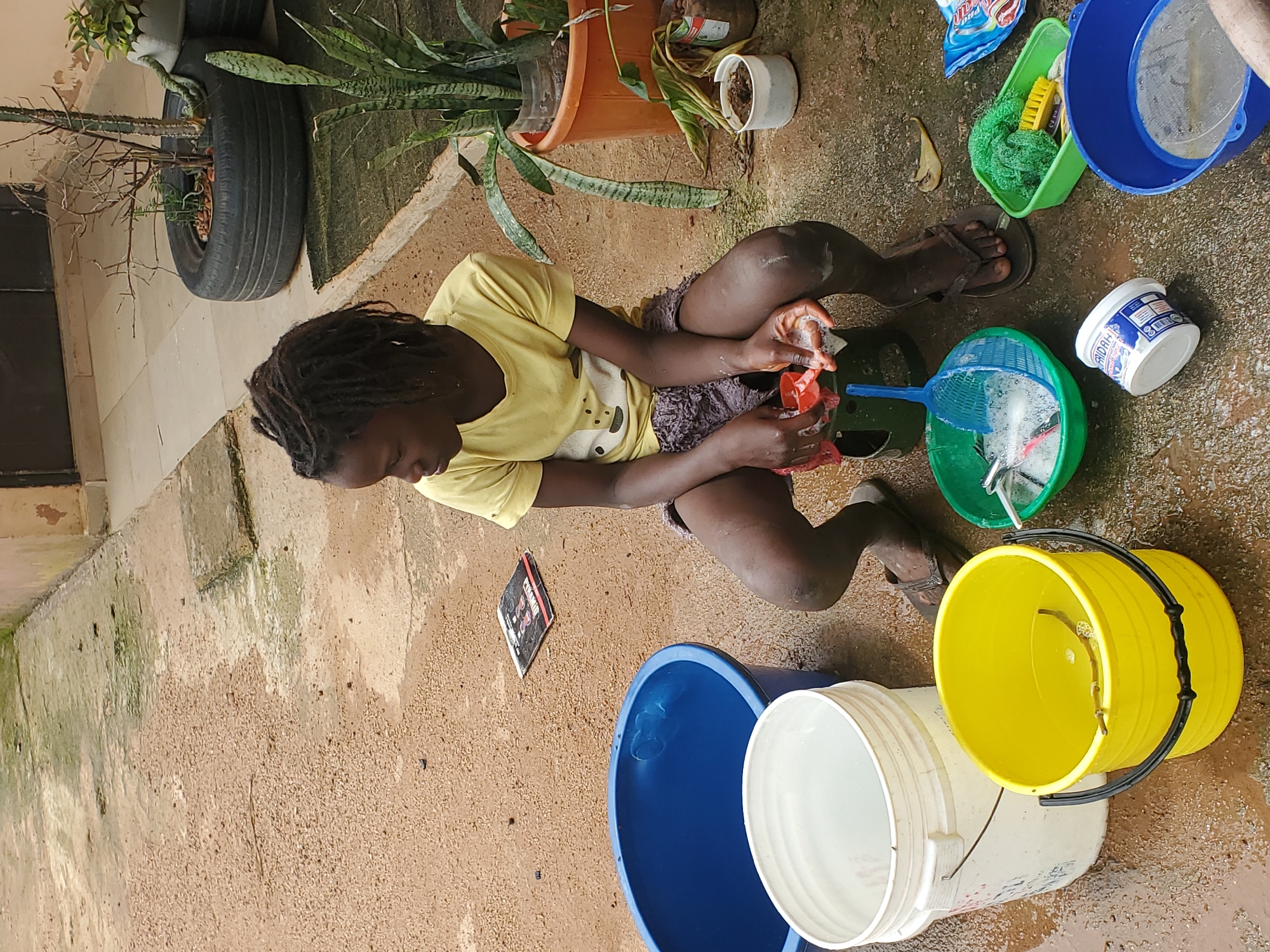 A young person with dreadlocks, wearing a yellow t-shirt and dark shorts, is squatting on a wet, sandy ground, engaged in washing. Their hands are soapy, and they are holding a small red object, possibly a toy or a cleaning tool, under a stream of water. Around them are various containers, including a large blue bucket, a yellow bucket, a white bucket, and a green basket containing a yellow brush and a green sponge. A blue colander with a mesh sieve is also visible, filled with soapy water. A blue and white detergent packet lies on the ground nearby.
In the background, there are plants, including a snake plant in a terracotta pot, and a tire repurposed as a planter with dry branches. The ground appears wet, suggesting recent rain or deliberate washing. The lighting and shadows indicate it is daytime. The overall scene suggests an outdoor domestic chore, possibly a wash day, taking place in a yard or communal outdoor space in Rafin Bauna, Nigeria. There is some visible text on the detergent packet, but it is not clearly legible. The person's posture and focused expression suggest concentration on the task at hand.