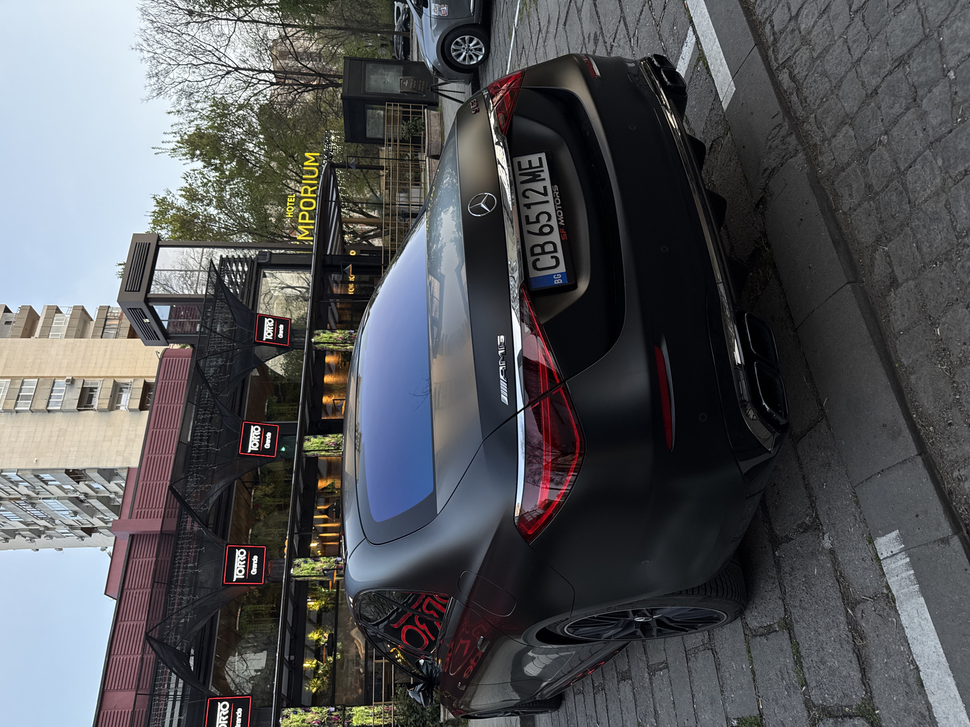 The image depicts the rear of a matte black Mercedes-AMG automobile parked on a cobblestone street, aligned with a painted white parking line. The license plate reads "CB 6512 ME," registered in Bulgaria ("BG"). The backdrop features a building with multiple "TORRO Grande" signs and the "HOTEL MPORIUM" sign above. Given the signs and architectural details, it is probable that the location is in the city of Plovdiv, Bulgaria. The car's position suggests it is parked next to the sidewalk.