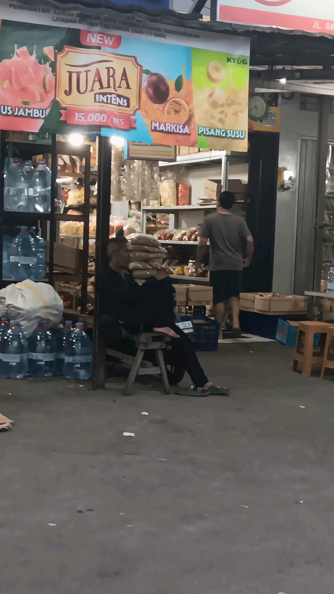The image captures a street scene in Depok, Indonesia, likely in the evening. The focal point is a stall with a large banner advertising juice drinks. The banner displays images of various fruits and labels like "JUARA INTENS," "MARKISA," and "PISANG SUSU." To the left of the banner, several large plastic bottles of water are stacked on shelves.
In the foreground, a man is seated on a small wooden stool, leaning against a shelf. He appears to be wearing dark clothing and sandals. Another man, seen from behind, is walking away from the stall towards the right side of the image. He is wearing a grey t-shirt and dark shorts.
The environment suggests a commercial area, possibly a market or roadside stall. Shelves behind the banner are stocked with various items, likely food products. Cardboard boxes are visible further in the background. The ground is paved and appears somewhat worn. Scattered small white objects, possibly litter, are present on the ground. The lighting suggests it is not daytime, with artificial lights illuminating the scene.