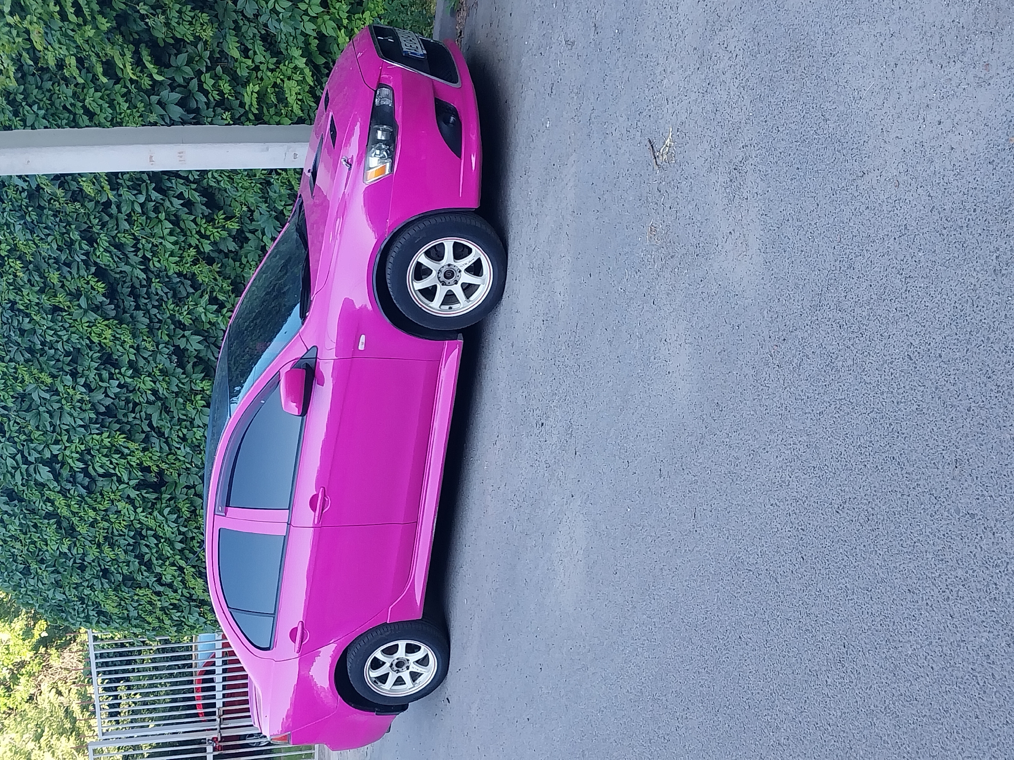 A bright pink Mitsubishi Lancer is parked on a paved surface in front of a dense green hedge. The car is angled, showing its side profile and the front passenger side. White multi-spoke wheels are visible on both the front and rear passenger side. The car has a black roof and dark-tinted windows. A concrete pillar is to the right of the car, and a metal fence is in the background to the left. The license plate of the car is partially visible. The scene appears to be an outdoor area in the city of Dnipro, Ukraine.