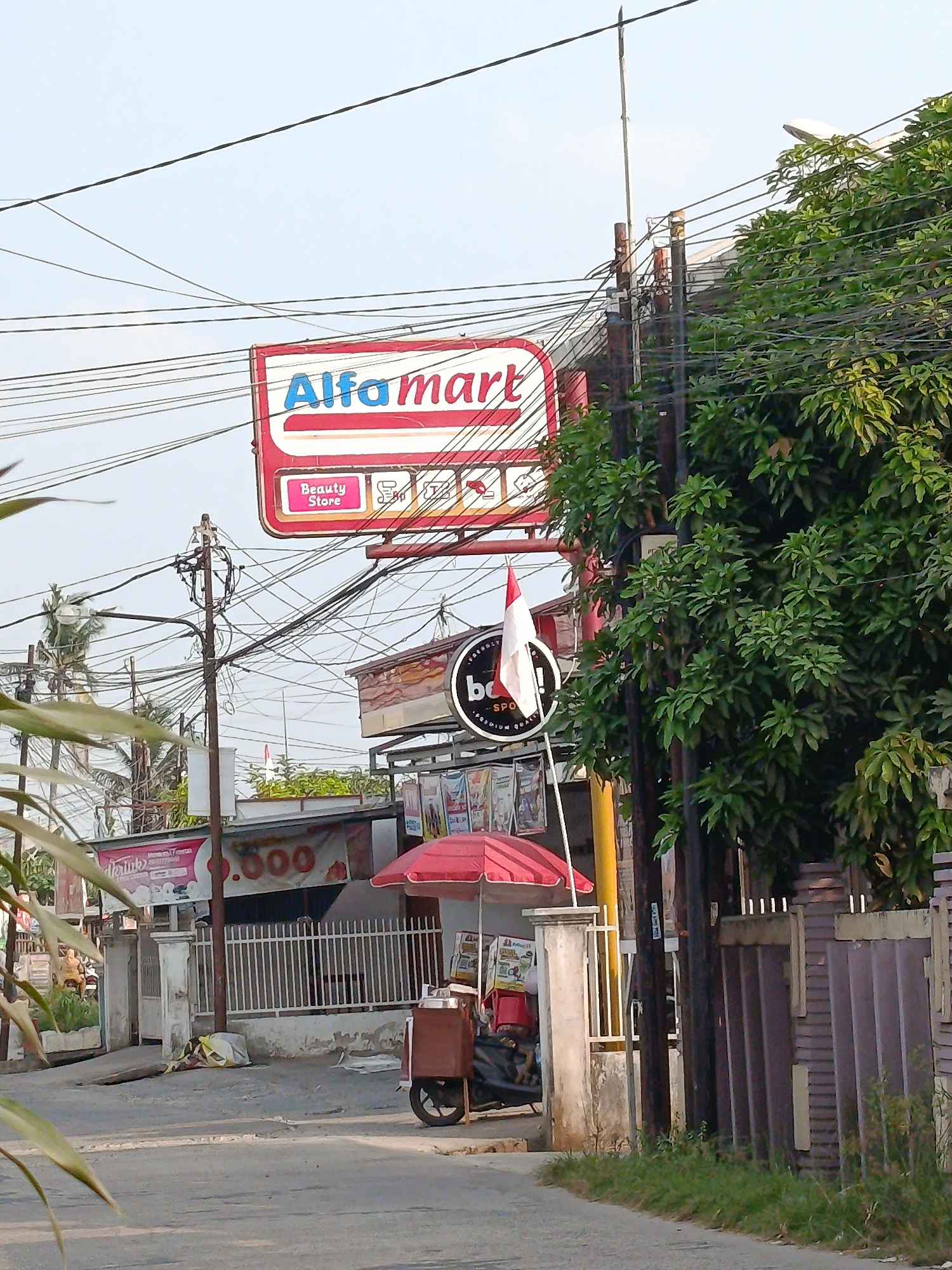 In Palembang, Indonesia, a street scene features a prominent Alfamart sign, a red and white building with a white fence, and a red umbrella covering a food cart. A person is seated on a motorcycle behind the food cart. The environment is a street with a sidewalk and a densely leaved tree. Overhead, a complex network of electrical wires crisscrosses the sky. A small red and white flag is visible on a pole near the food cart. The buildings appear to be shops, with some advertising banners visible. The road surface is asphalt.