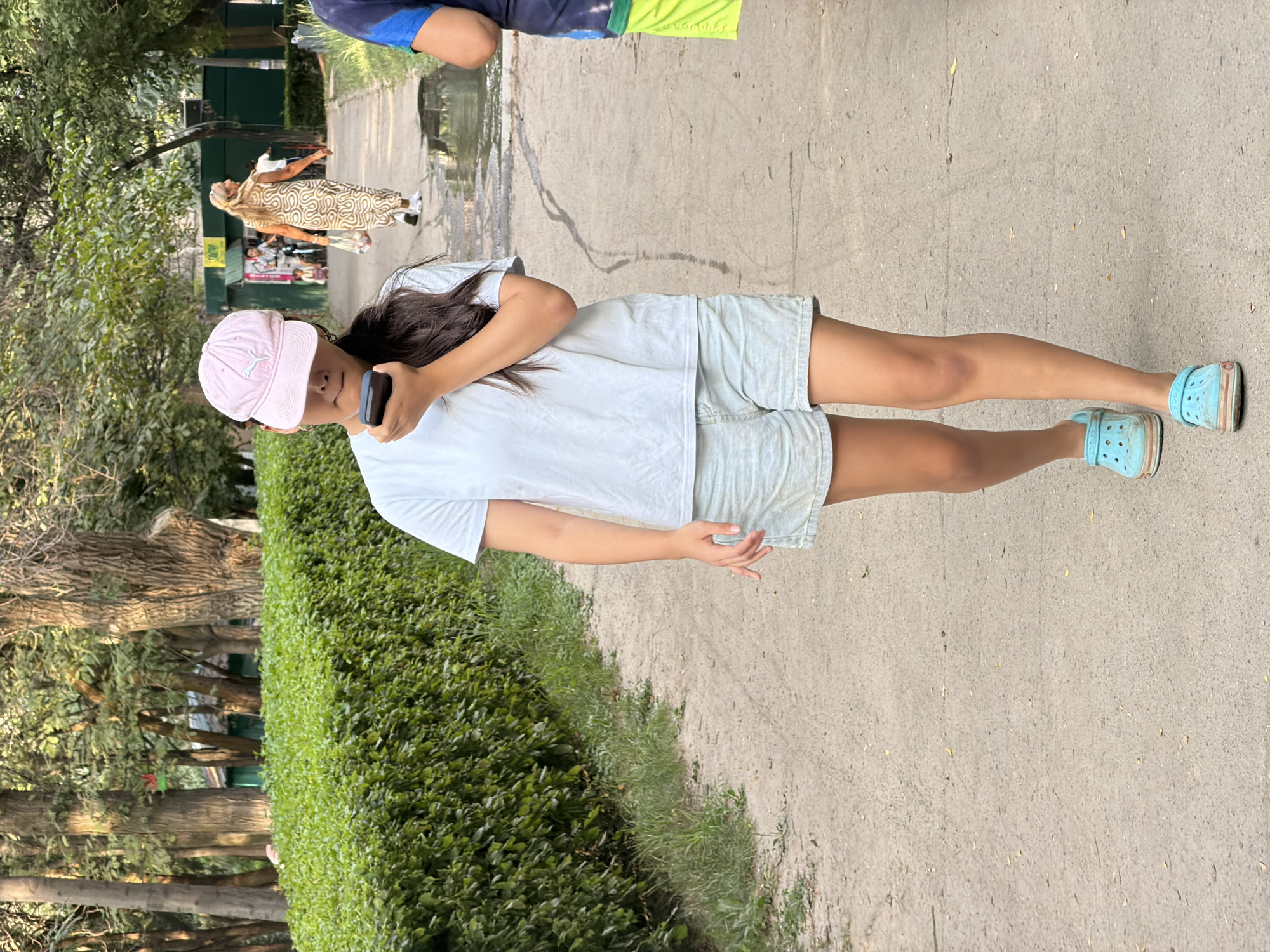 In an outdoor park setting, a young woman walks forward on a dirt path, holding a dark-colored mobile phone to her right ear. She is wearing a light blue t-shirt, light blue denim shorts, a pink baseball cap, and light blue clogs. Her dark hair is pulled back. A green hedge runs along the left side of the path, and trees with green foliage form a backdrop on the left. In the background on the right, a woman in a patterned dress walks away from the camera, and other people are visible further down the path. The ground appears wet in places.