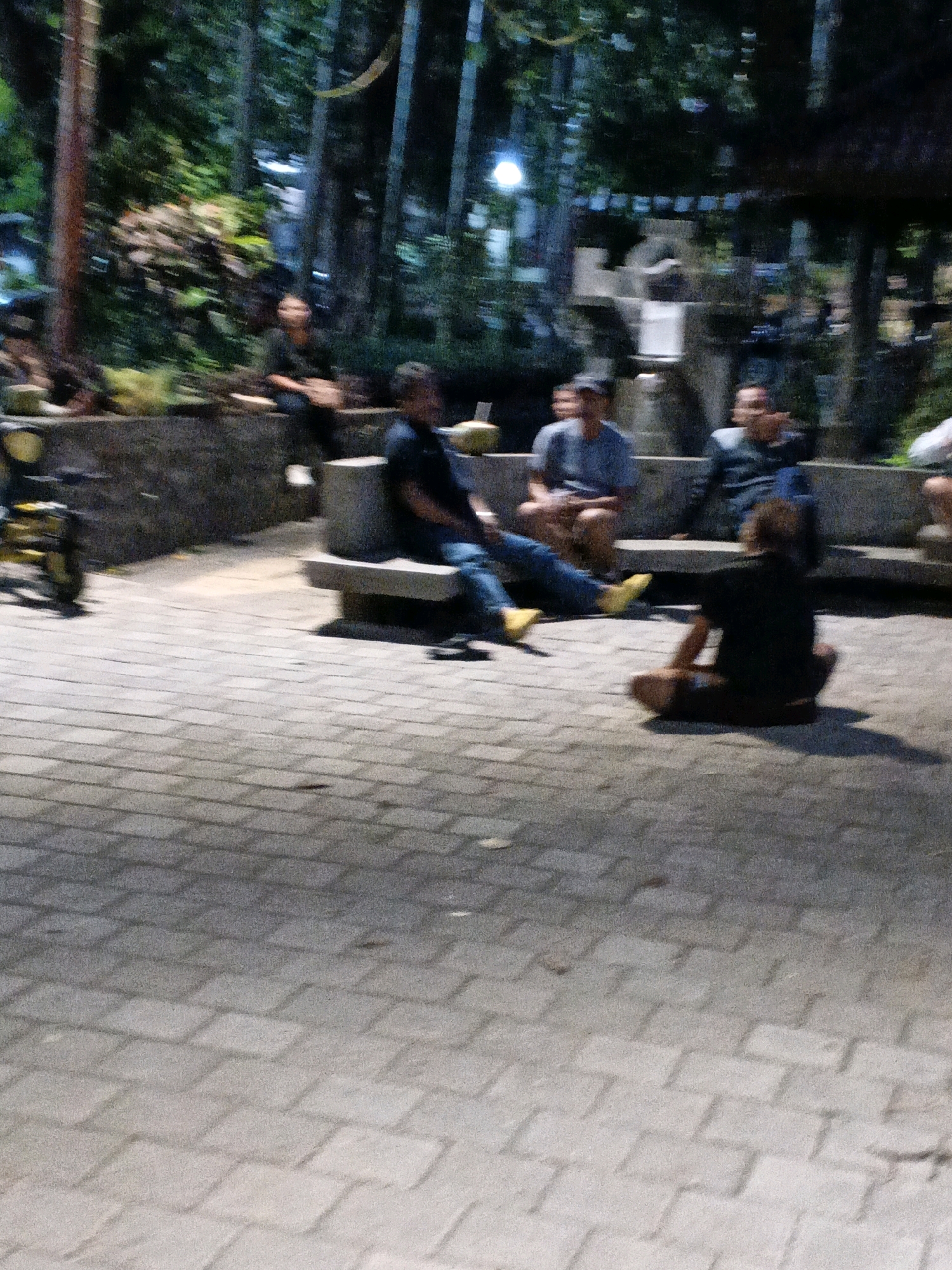 A group of people are sitting and standing in what appears to be a park or outdoor seating area at night. The ground is paved with interlocking bricks, and there are benches and some landscaping visible in the background. The lighting is dim, creating shadows. One person is sitting cross-legged on the ground. Others are seated on benches. The location is identified as Denpasar, Indonesia.